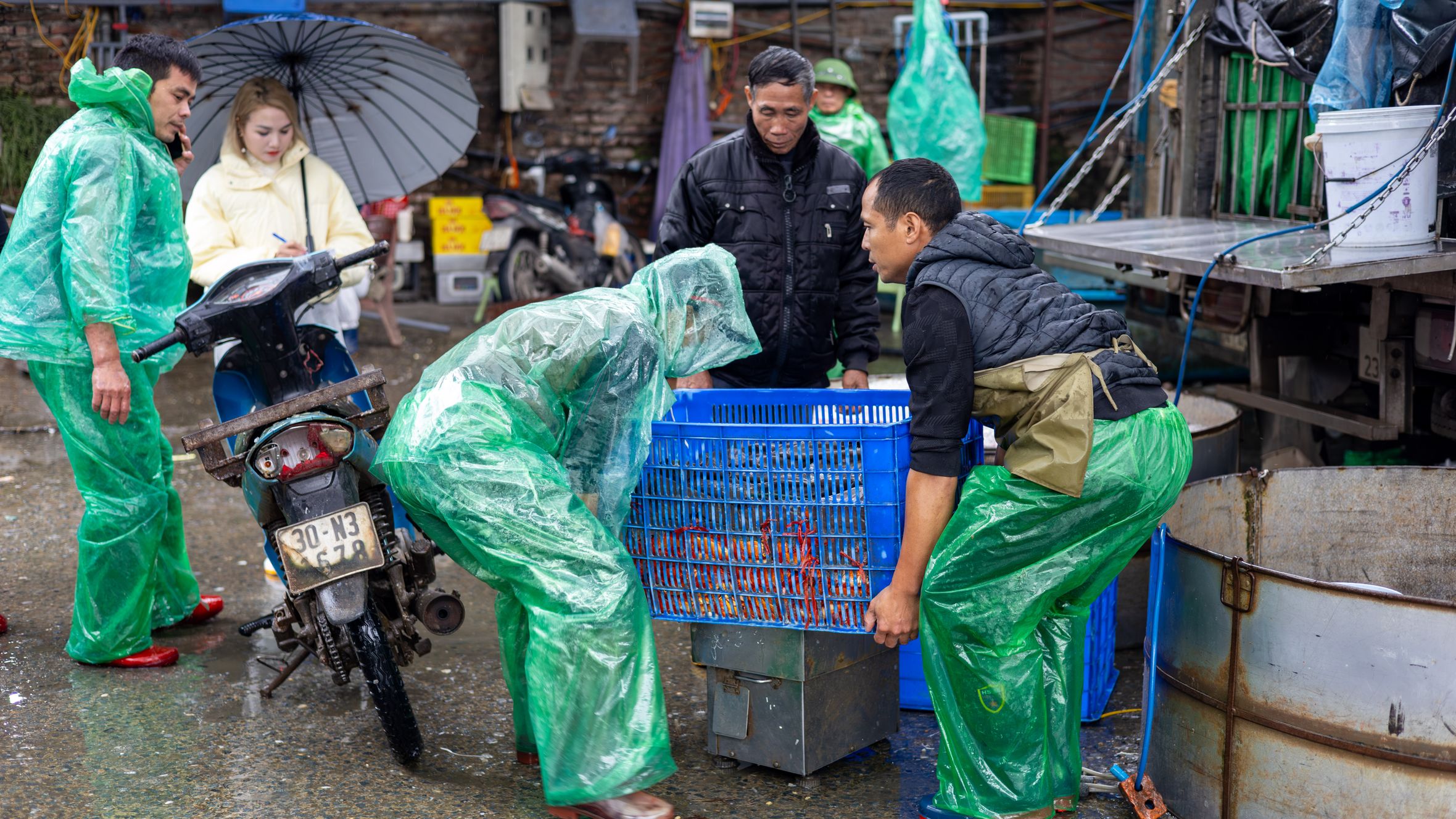 Workers in raincoats managing crates on a bustling street in Hanoi during wet weather.