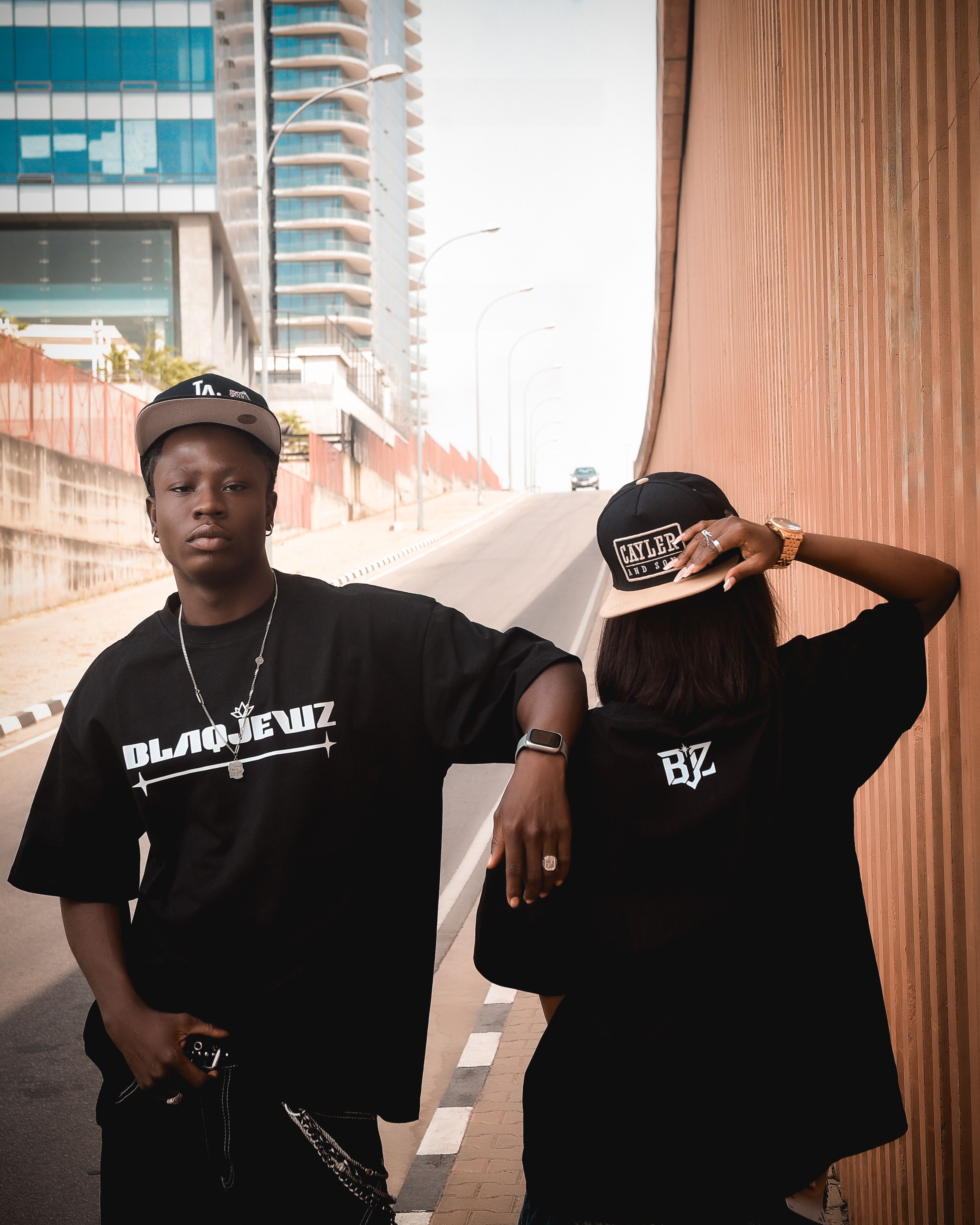Two young adults in matching black streetwear pose confidently in urban setting.