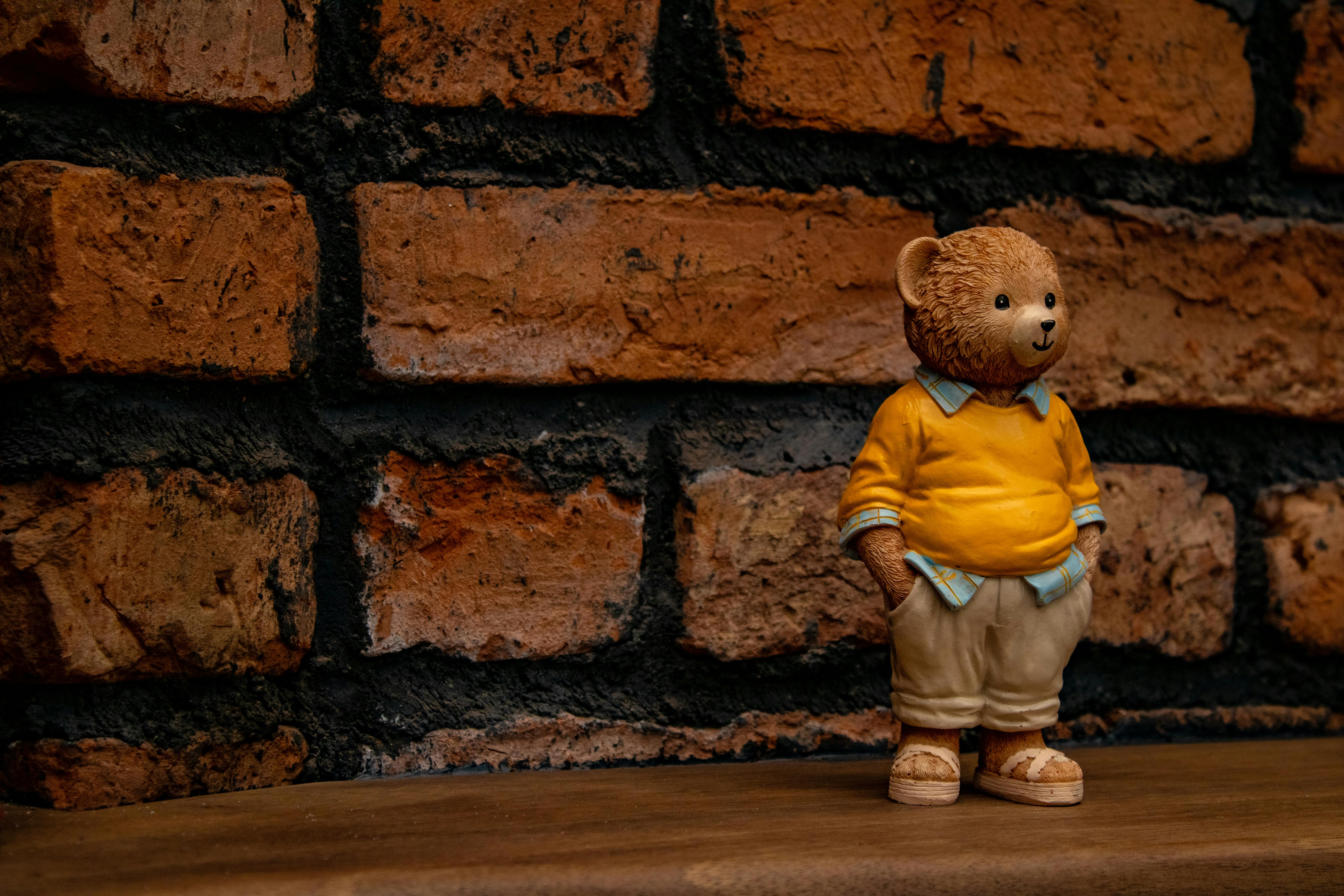免费 可爱的泰迪熊摆件，身穿黄色衣服，倚靠在古朴的砖墙上。非常适合温馨的家居装饰。 素材图片