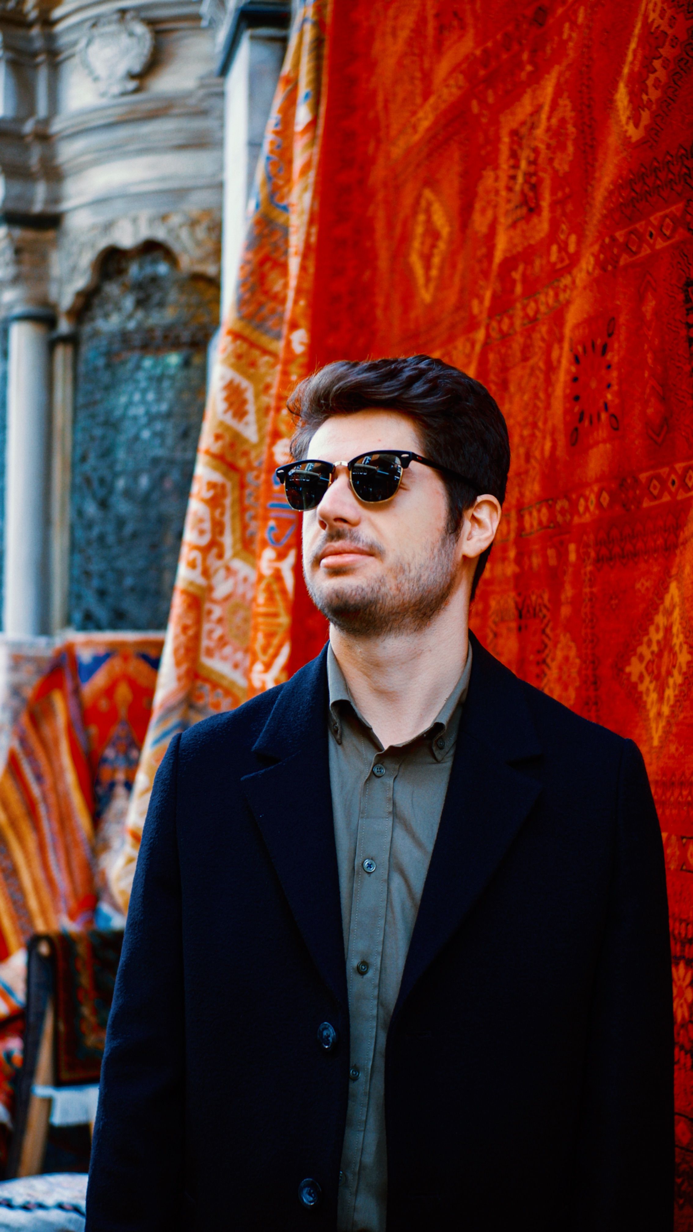 Stylish man in sunglasses stands by vibrant Turkish rugs in Istanbul, showcasing cultural elegance.