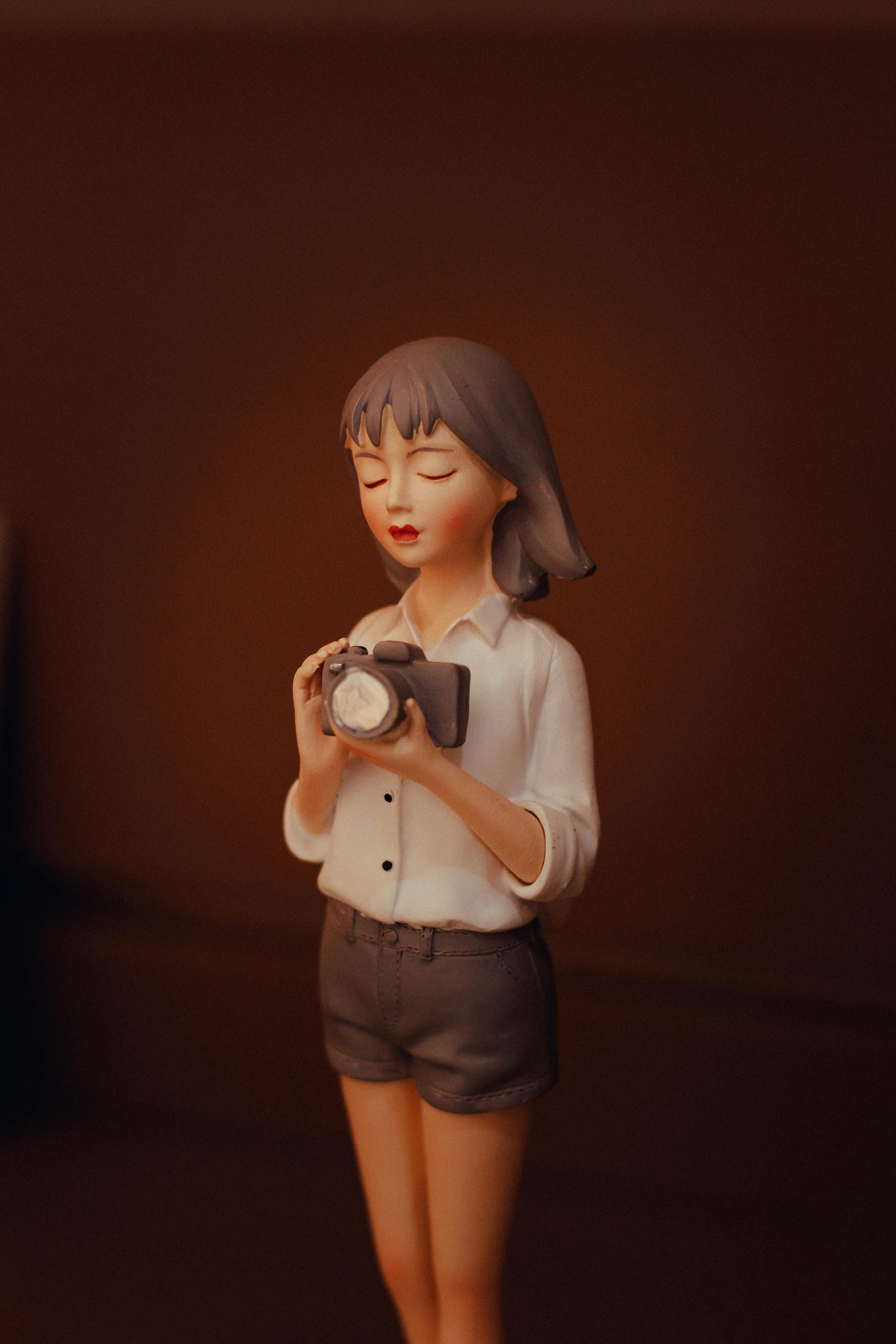 Free A whimsical figurine of a girl with closed eyes holding a camera, set against a warm brown backdrop. Stock Photo