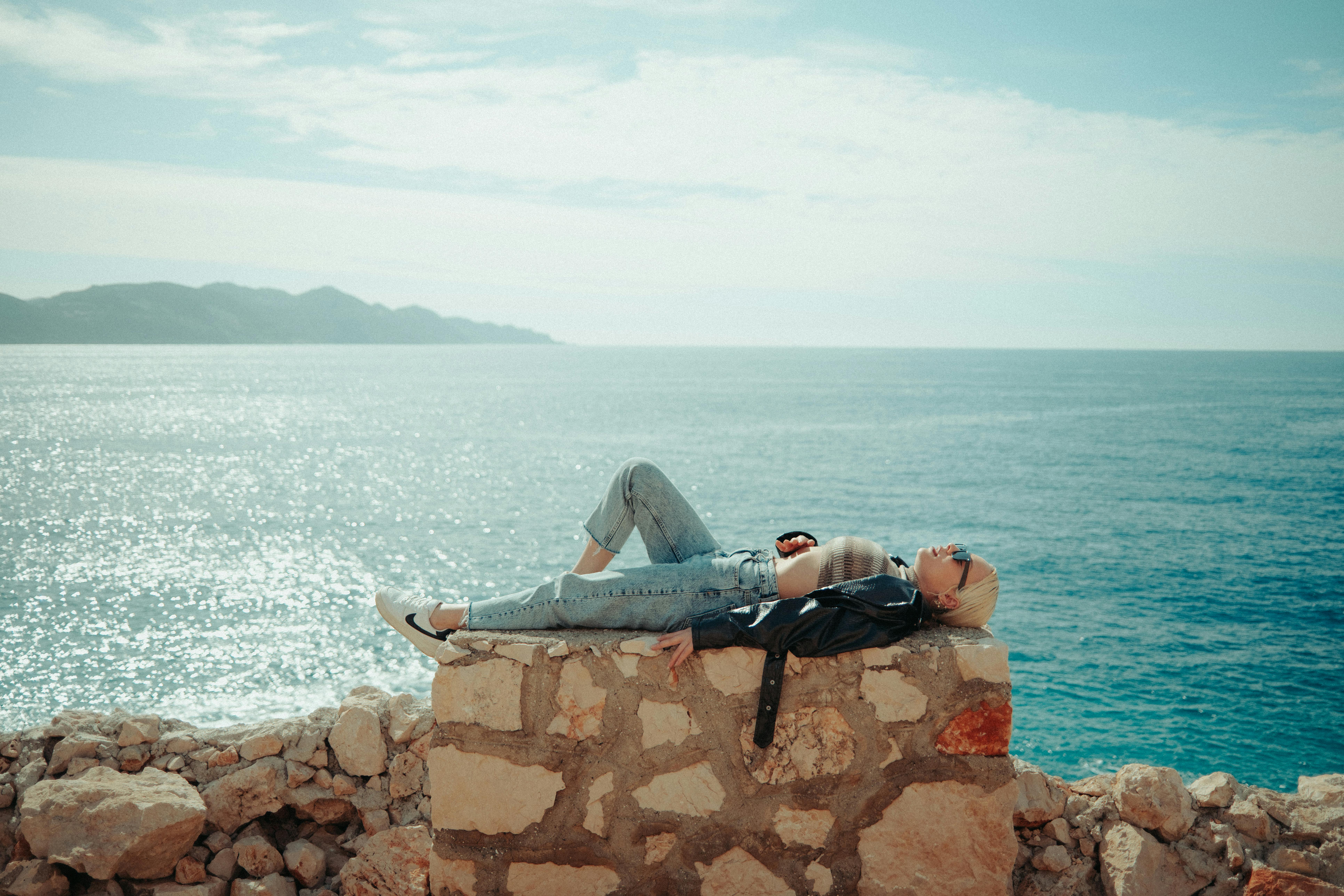 Безкоштовне Жінка в повсякденному одязі відпочиває на кам'яній стіні біля блискучого моря сонячного дня. стокове фото