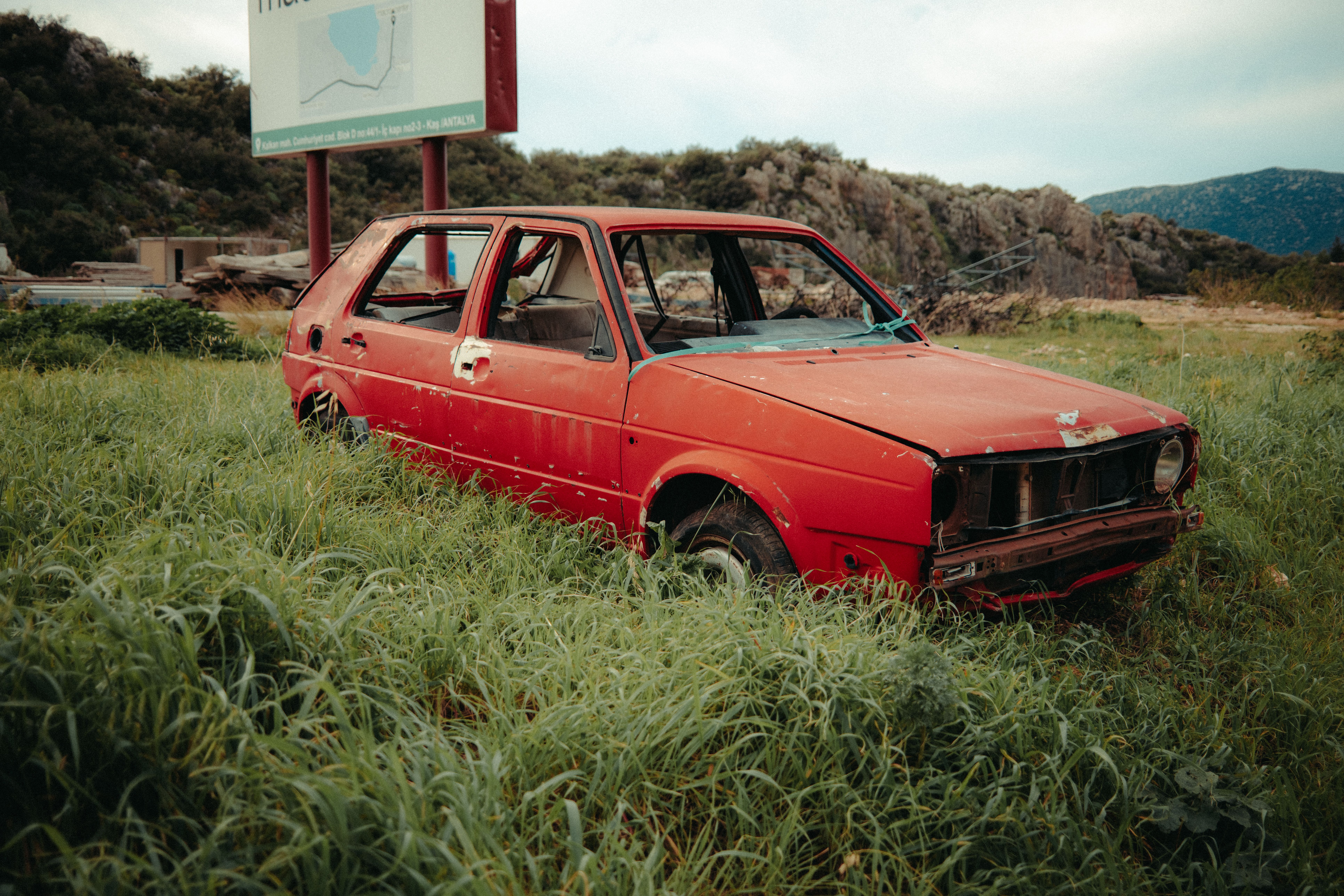 An old, rusting red car abandoned in a lush, overgrown grassy field under a cloudy sky.