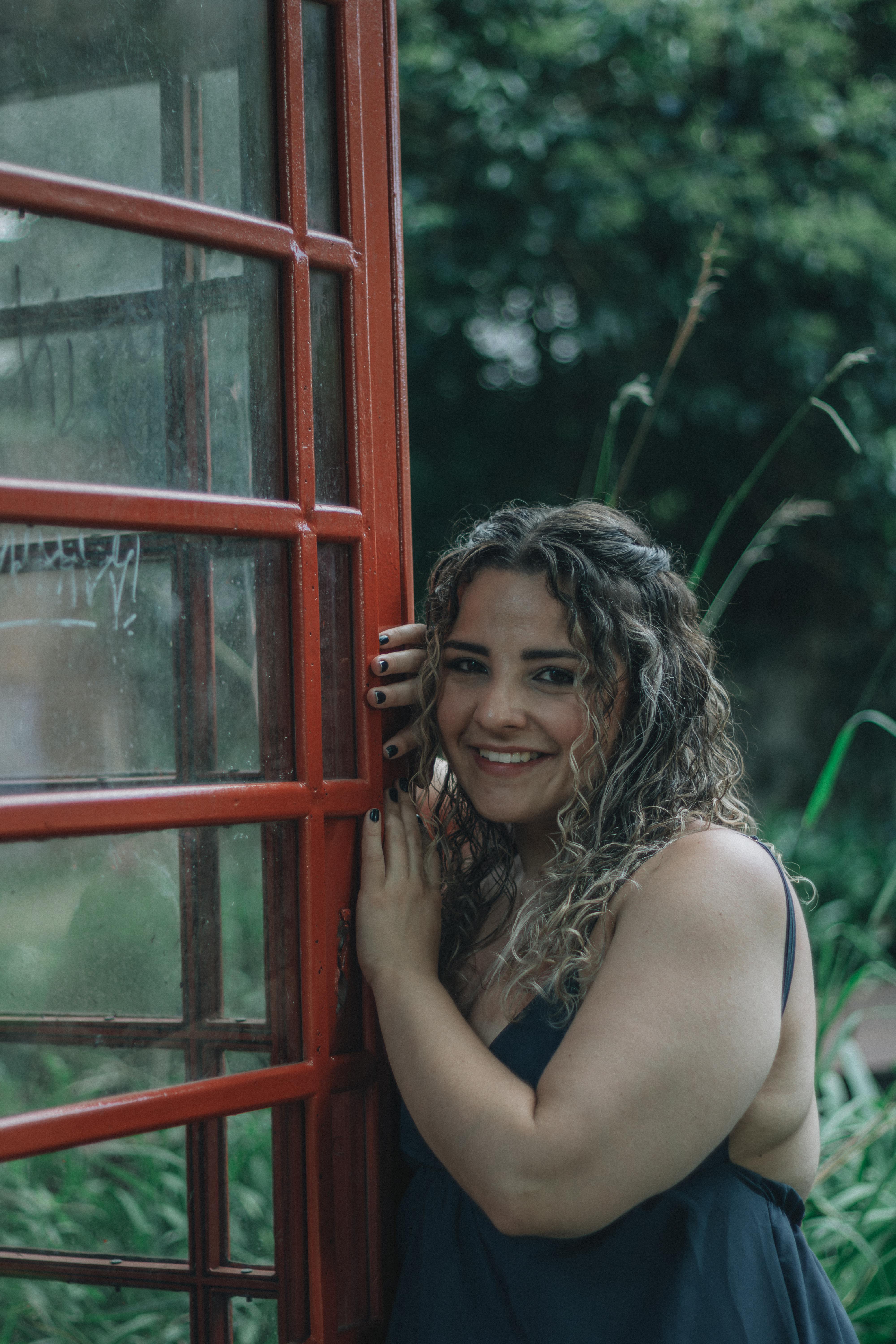 ฟรี หญิงสาวผู้เปี่ยมด้วยความสุขกำลังโพสท่าอยู่ข้างตู้โทรศัพท์สีแดงแบบดั้งเดิม ท่ามกลางสวนอันเขียวชอุ่ม คลังภาพถ่าย