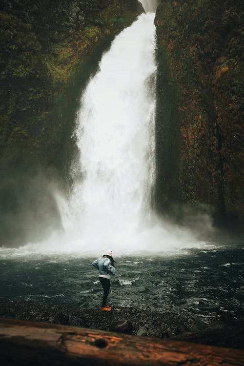 Person in Blue Jacket and Black Pants Standing on Rock Near Waterfalls