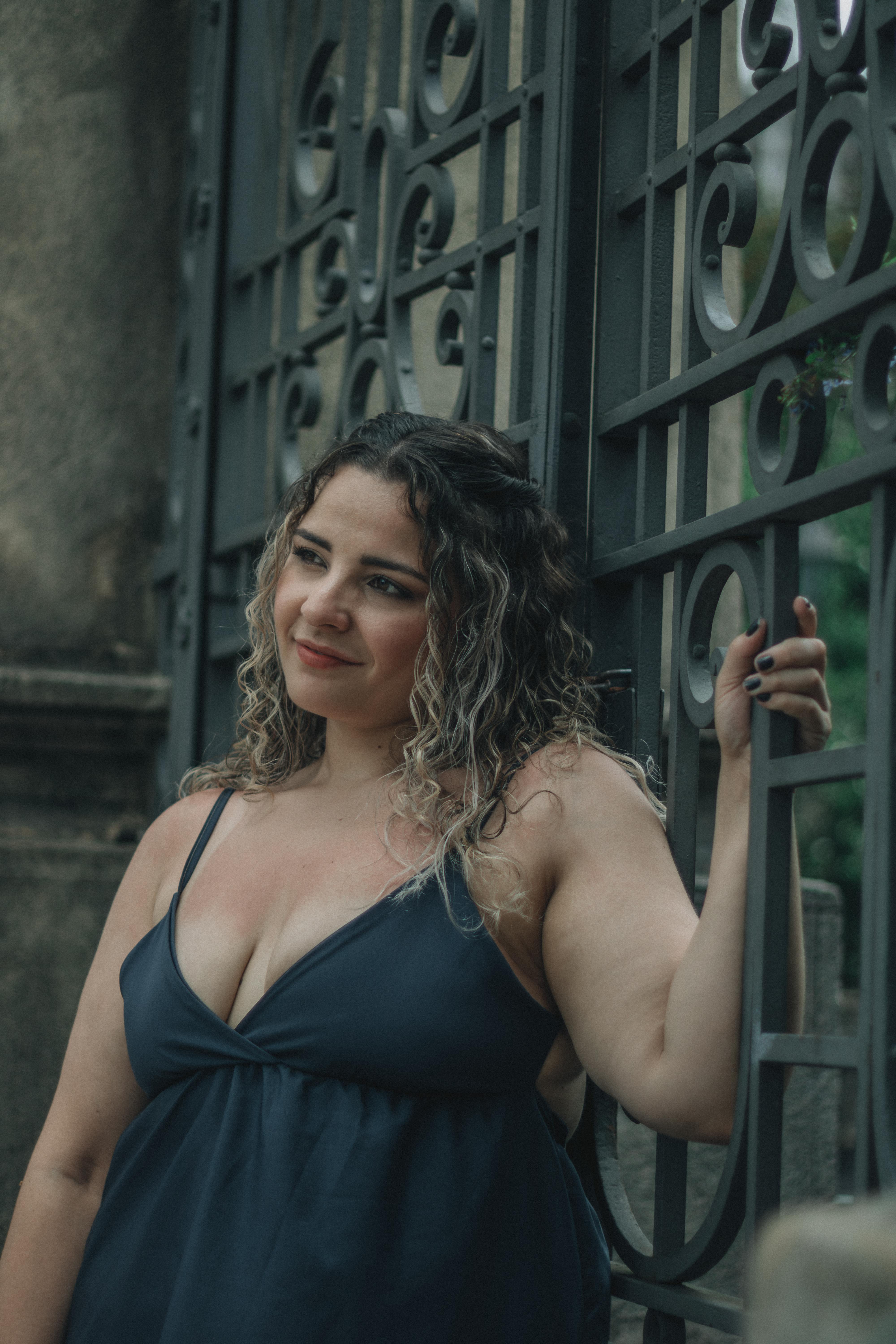 Kostnadsfria Lockigt hårigt kvinna i blå klänning poserar vid en dekorativ grind utomhus. Stock foto