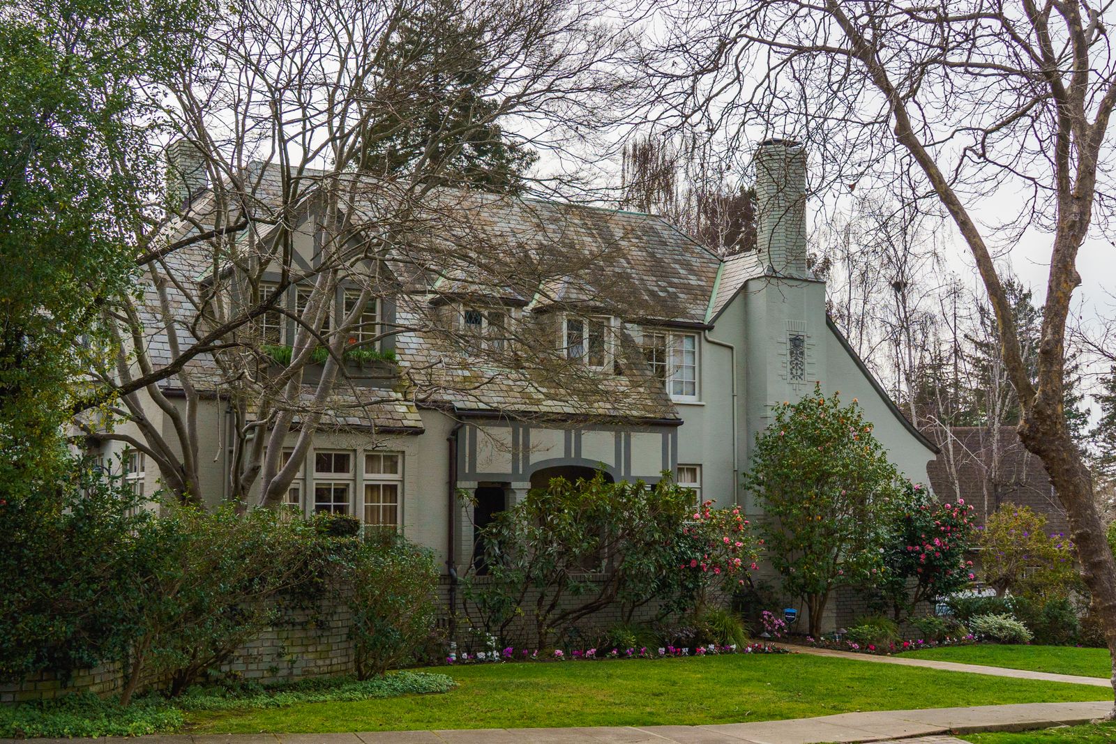Tudor-style home with a steep gabled roof, half-timbered stucco facade, tall brick chimney, and leaded windows, framed by mature trees and flowering shrubs, Royce Woods, Naperville IL