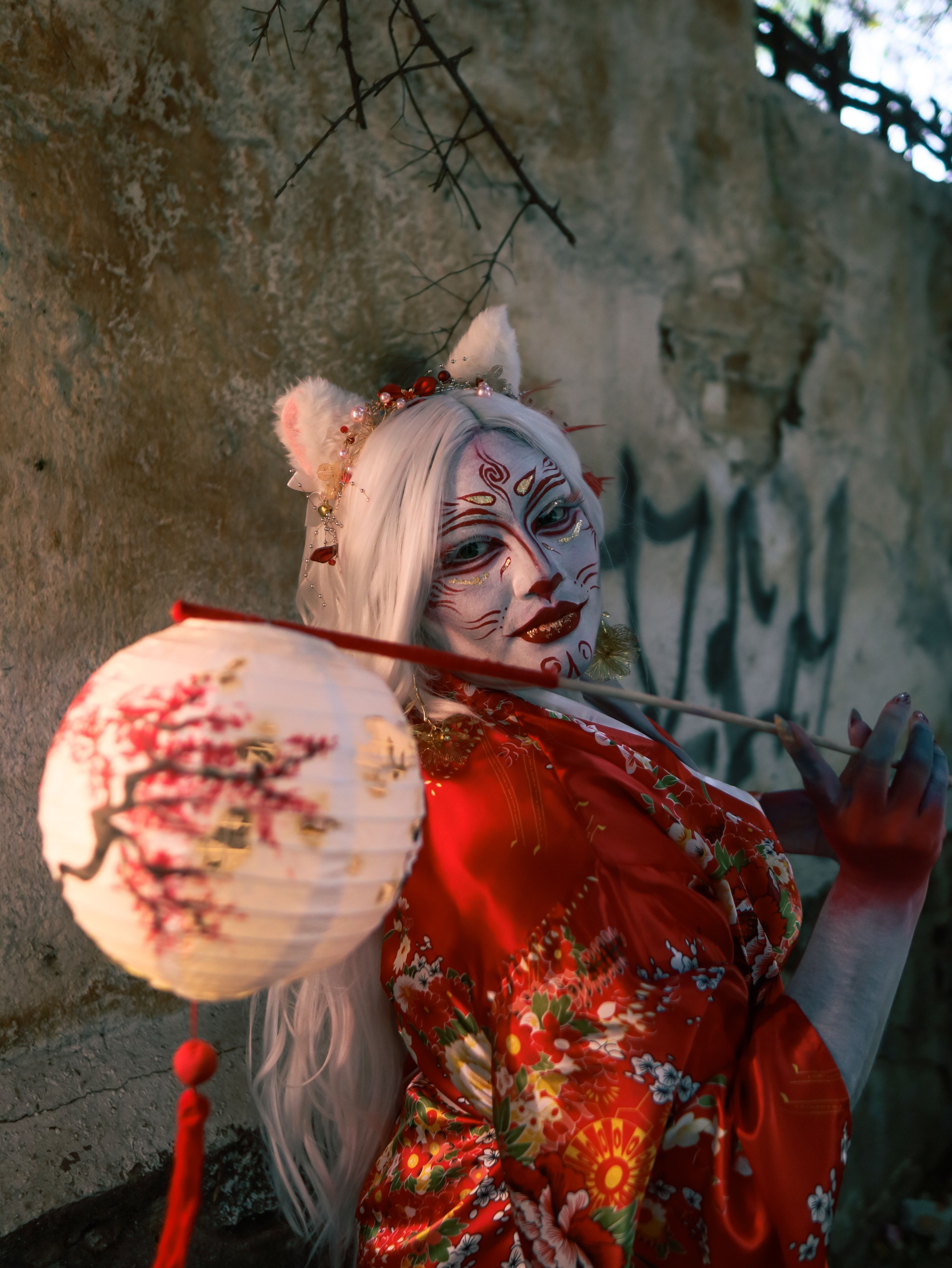 A woman in a vibrant red kimono with artistic face paint holding a lantern in a moody setting.