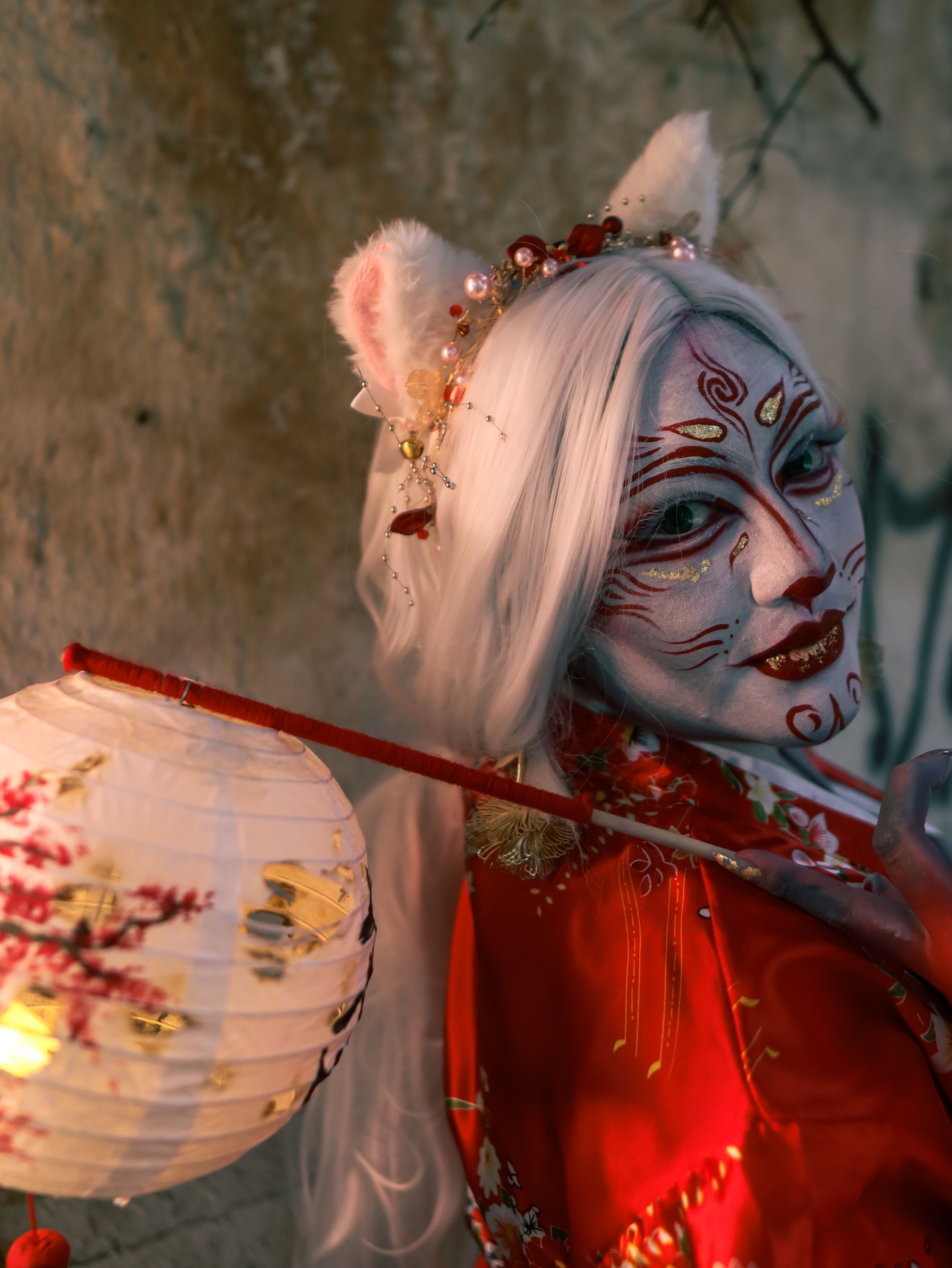 Enchanting kitsune-inspired performer in vibrant red kimono with face paint, holding a paper lantern.
