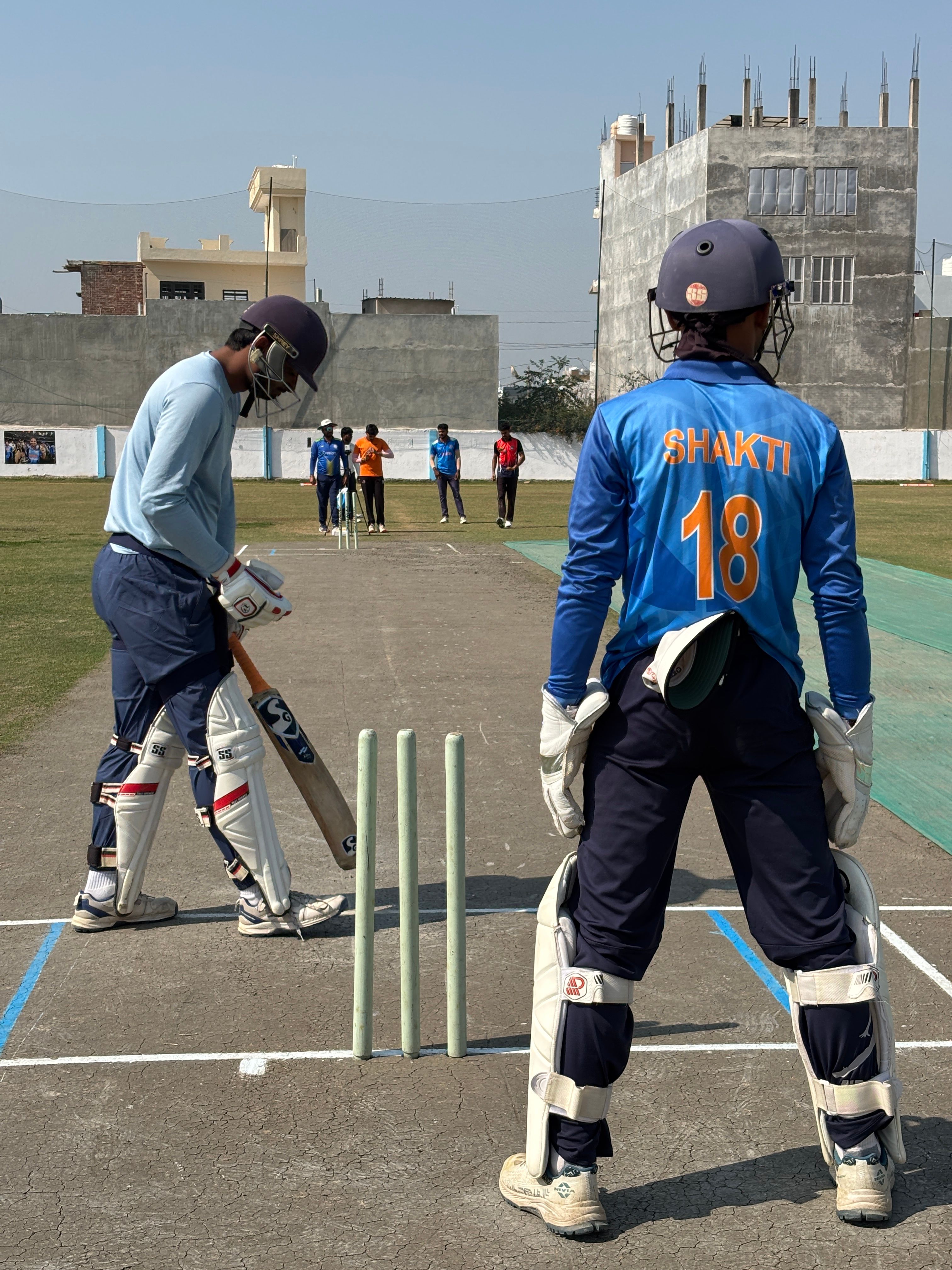 Players practicing cricket on an outdoor ground under clear skies.