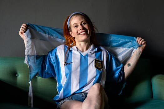A woman joyfully wearing an Argentina soccer jersey while sitting indoors. Celebrating with national pride.