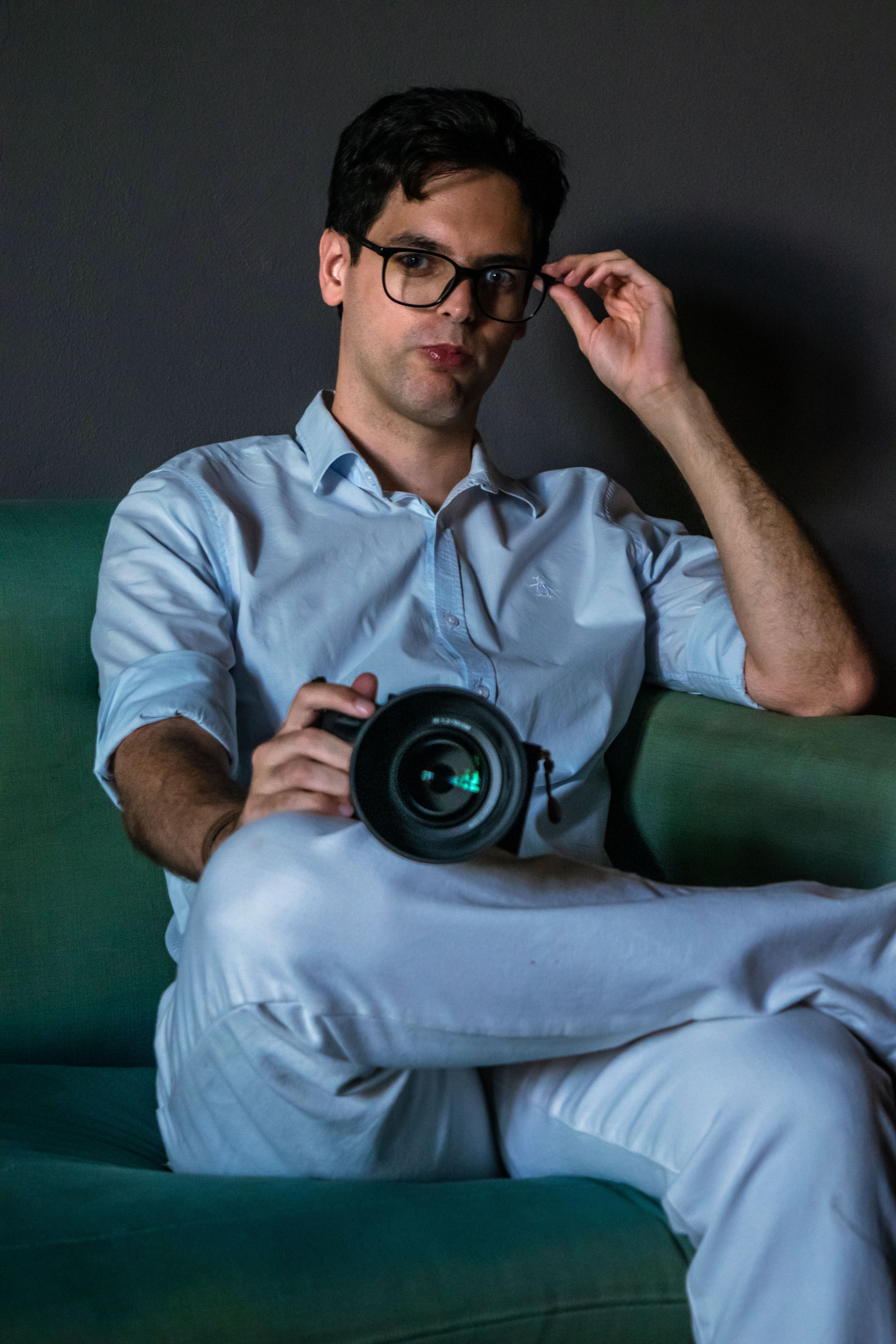 Man posing with camera on a green sofa, adjusting glasses indoors.