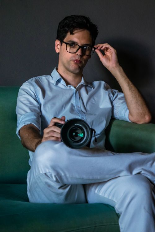 Free Man posing with camera on a green sofa, adjusting glasses indoors. Stock Photo