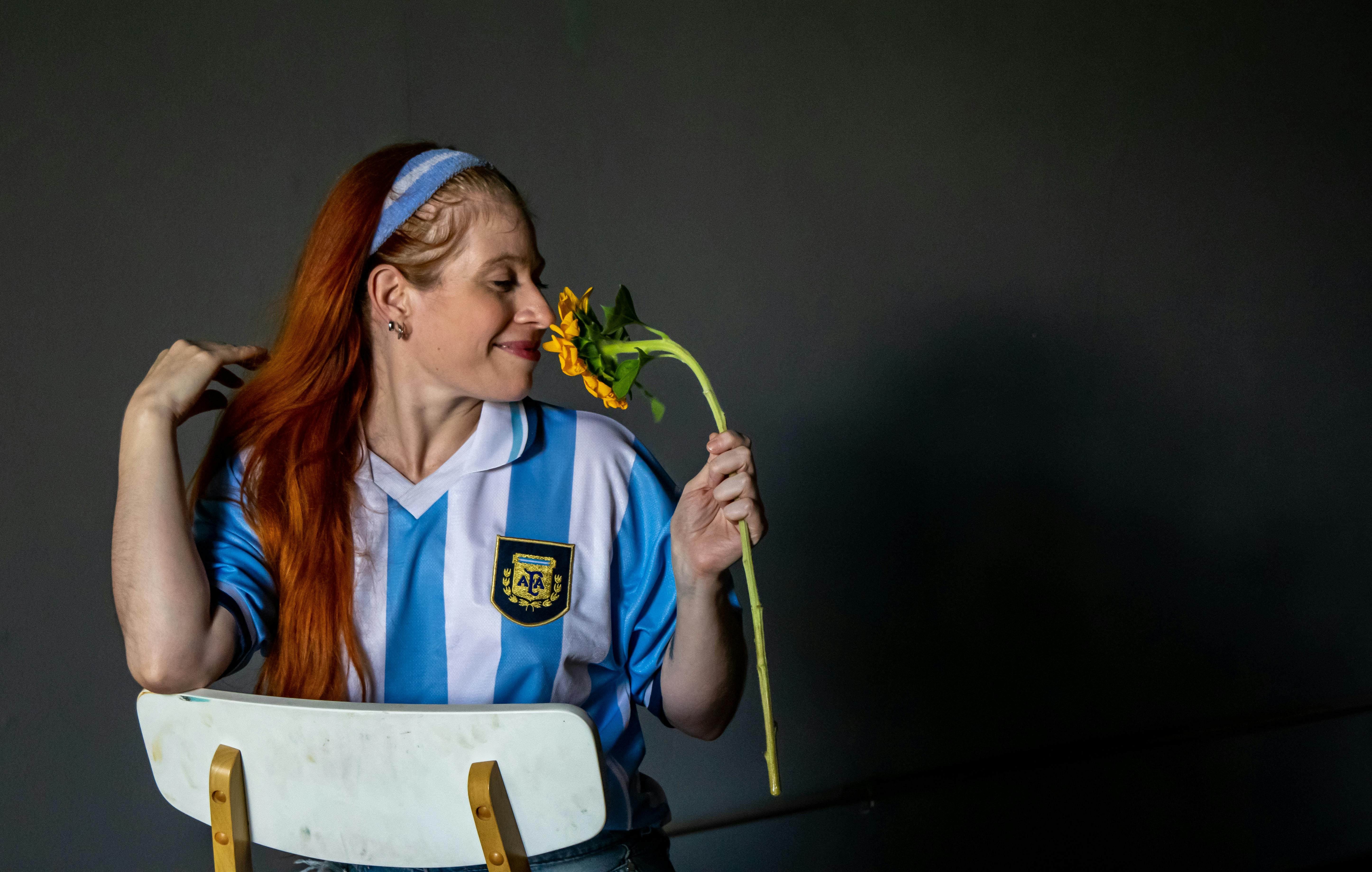 бесплатная Рыжеволосая женщина в сине-белой полосатой футболке наслаждается ароматом жёлтого цветка в помещении. Стоковое фото