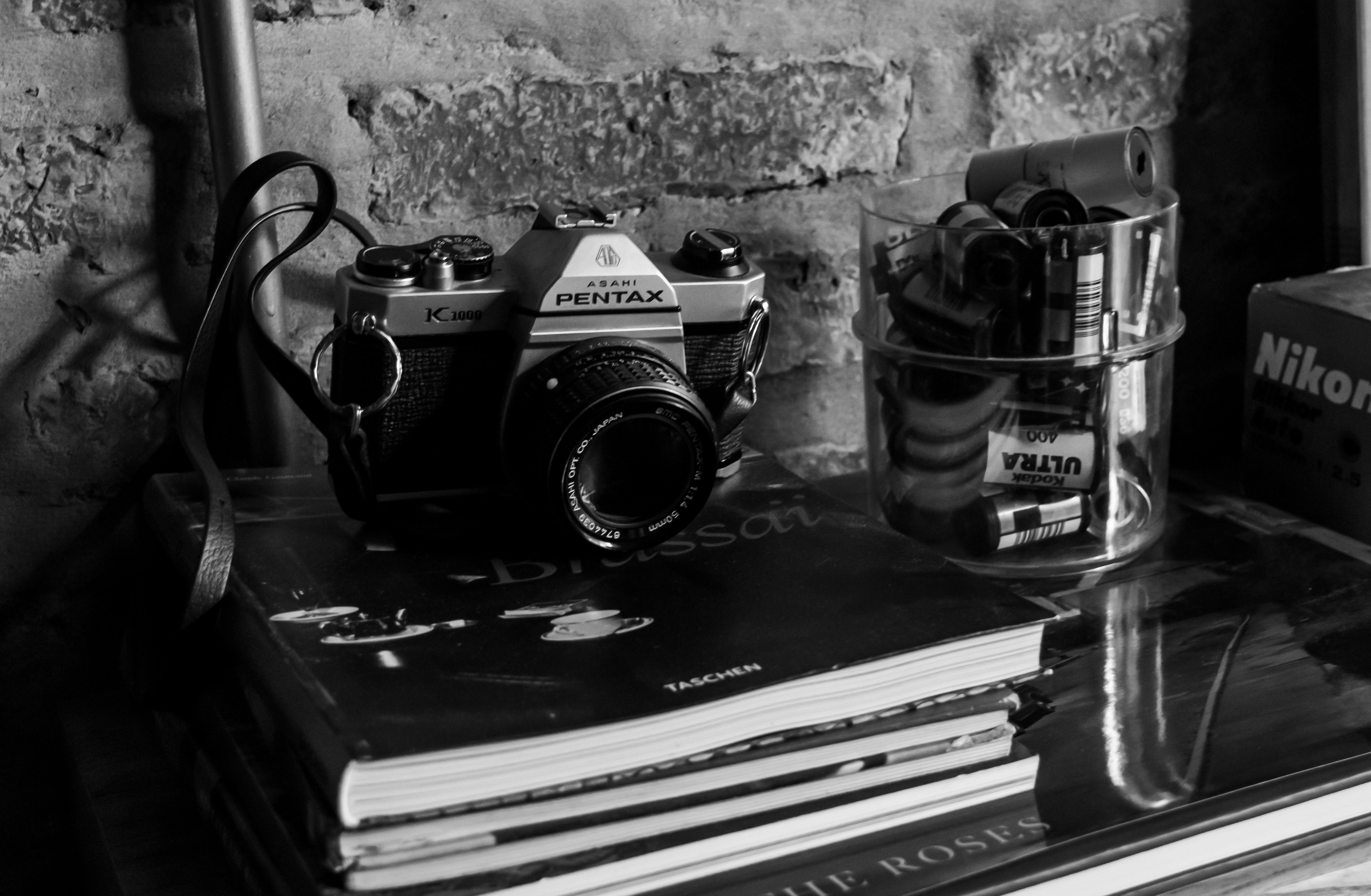 Free Black and white photo of a vintage Pentax camera on a shelf with film and books. Stock Photo