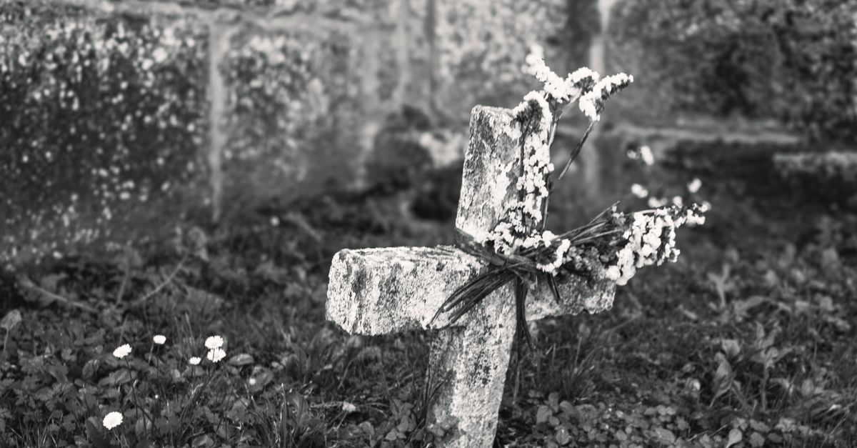 A black and white photo of an old stone cross adorned with flowers in a cemetery setting.