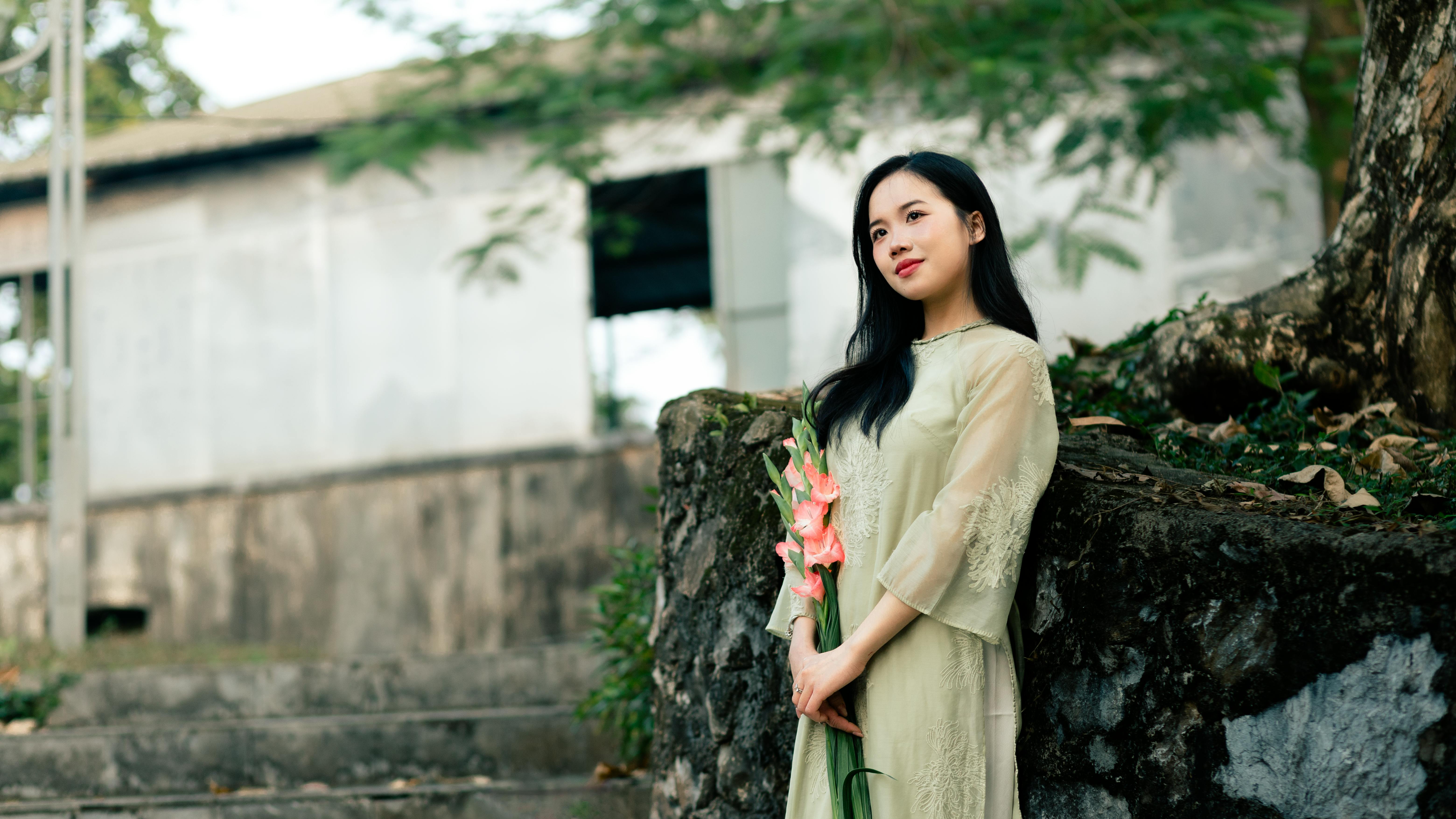 Gratis Una donna serena con un lungo abito verde chiaro tiene in mano dei fiori, all'aperto, vicino a una vecchia struttura. Foto a disposizione