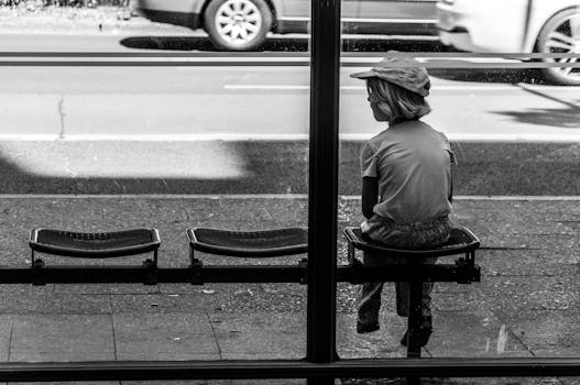 A black and white photo of a child sitting alone at a bus stop on an urban street.