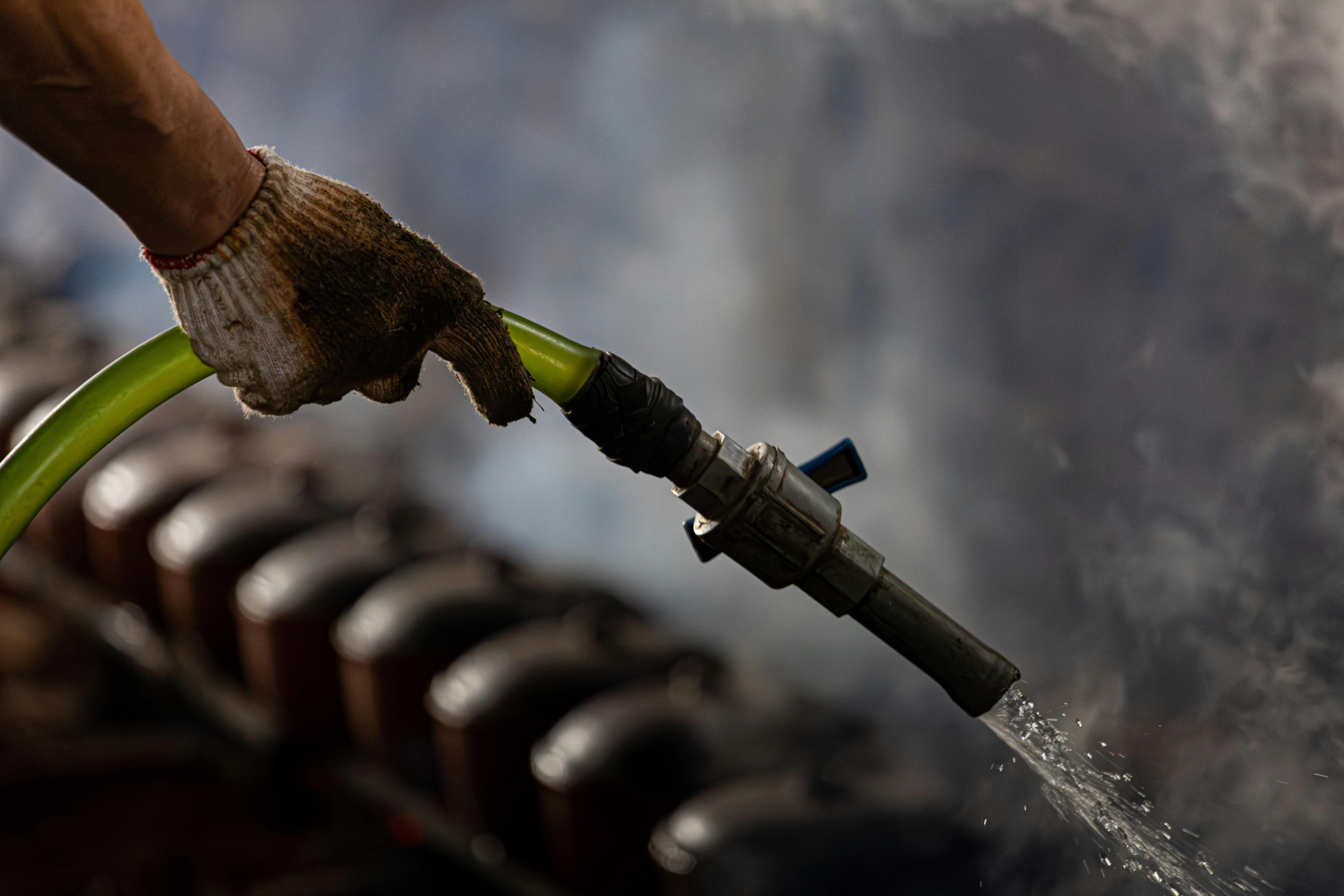 Gratis Una mano enguantada manejando una manguera de agua industrial con vapor en el fondo. Foto de stock
