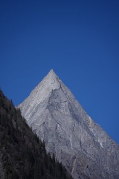 A sharp, rocky mountain peak with lush green forest under a clear blue sky captures nature's beauty.