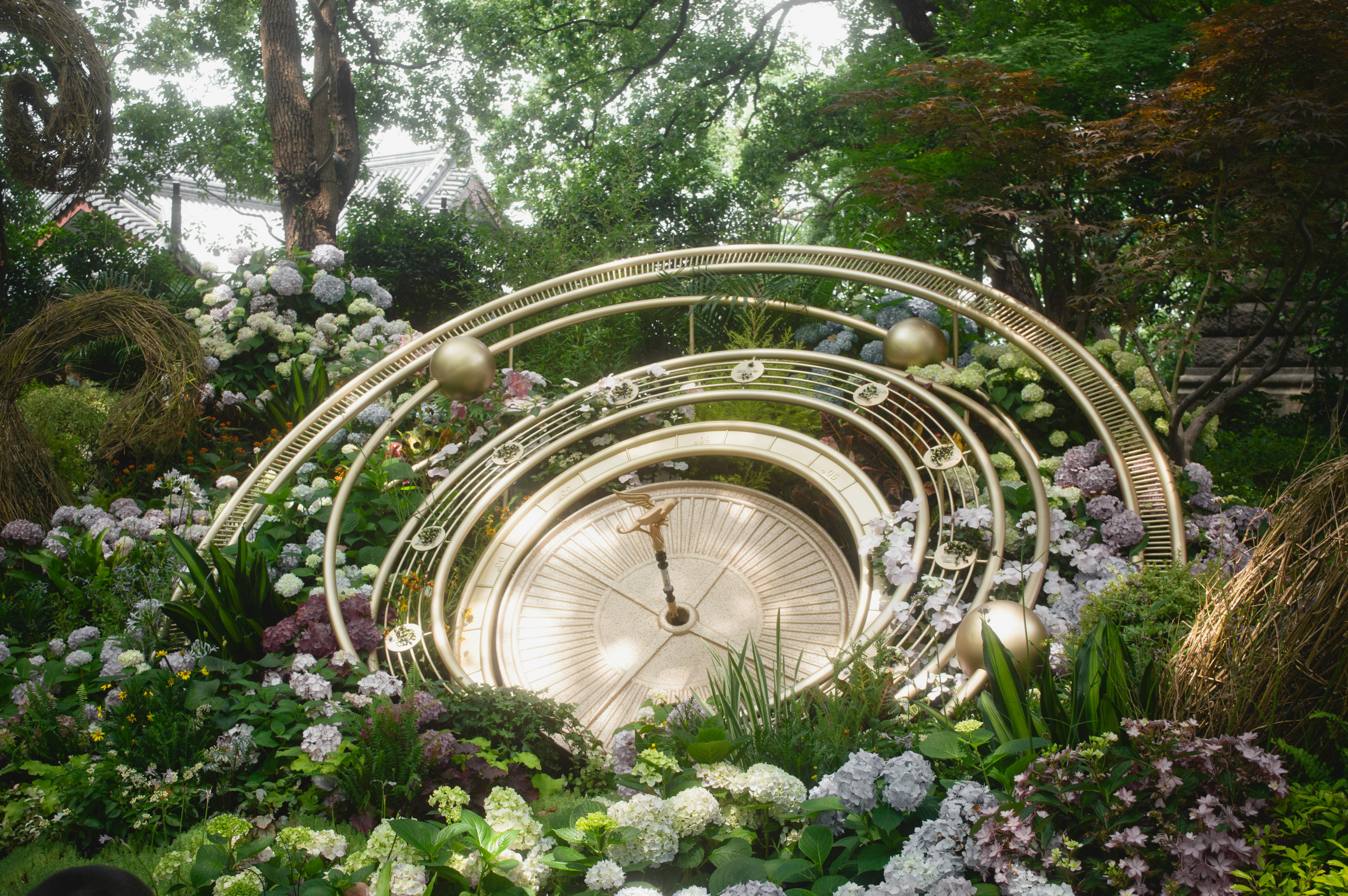 Kostenlos Eine große Zieruhr steht inmitten üppiger grüner Pflanzen und Blumen und schafft so eine ruhige Gartenszene. Stock-Foto