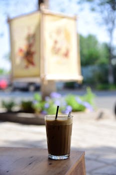 A glass of iced coffee with a straw on a wooden table outdoors, sunny day.