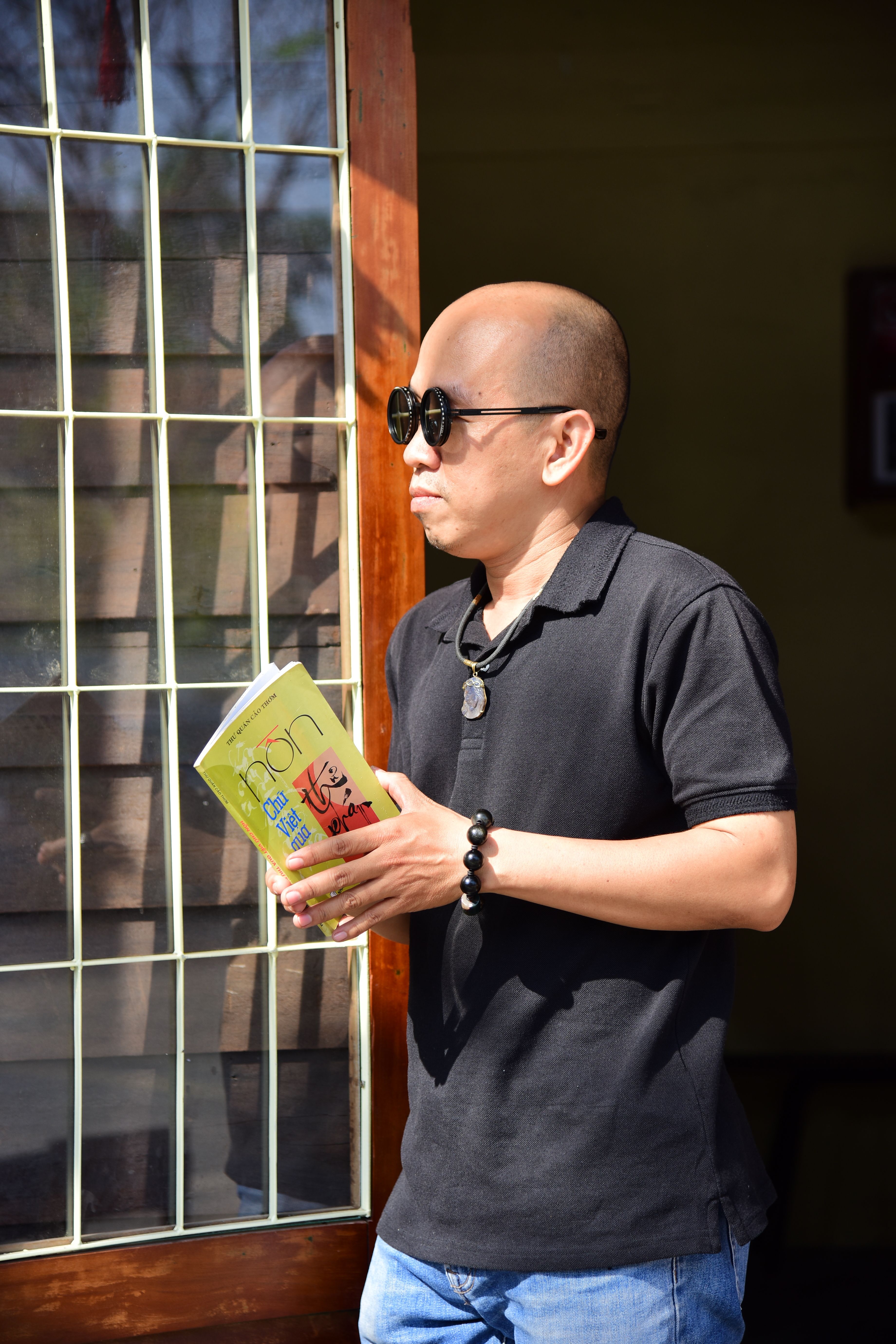 A man holding a book stands by a sunlit window, wearing sunglasses and casual attire.