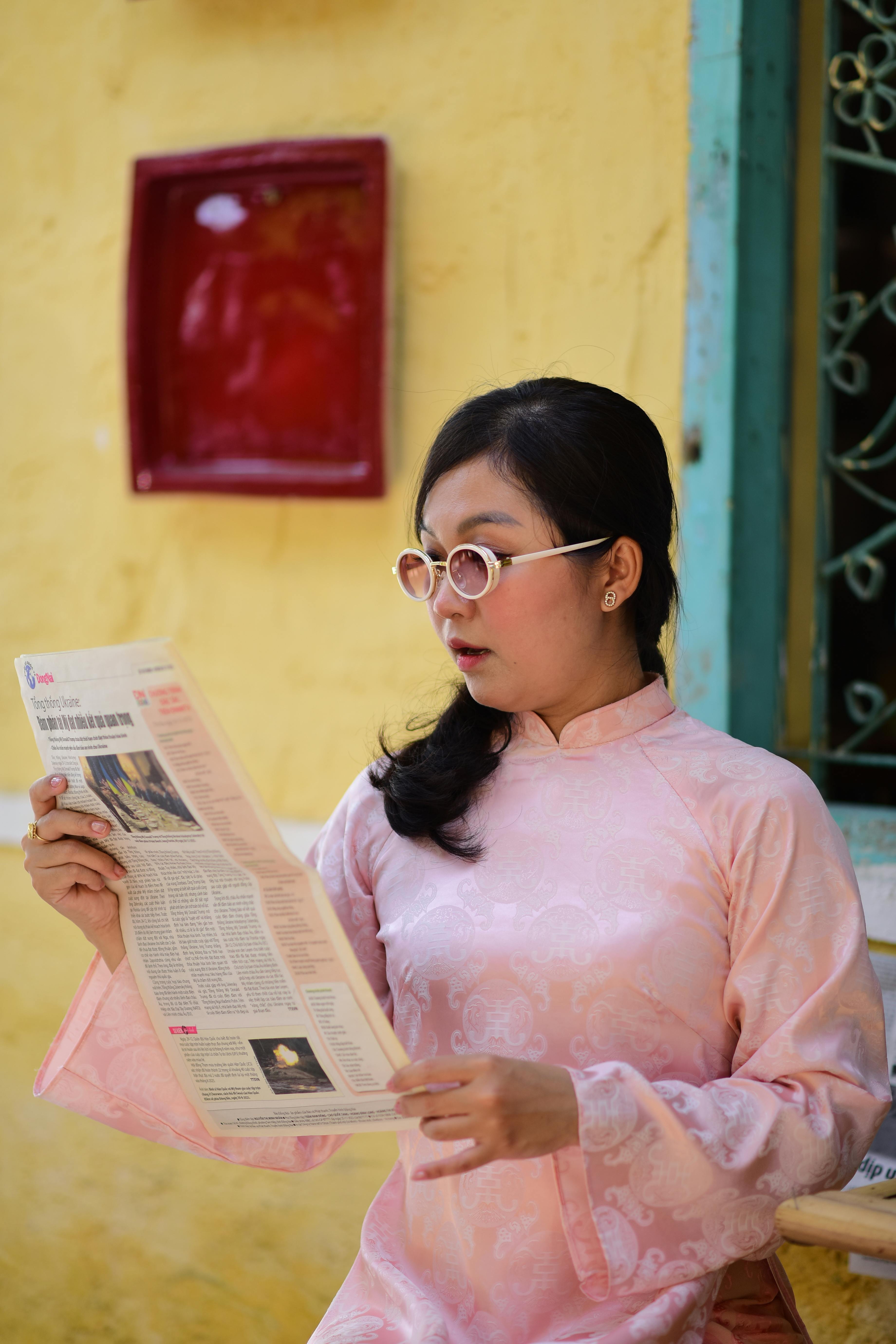 Gratis Donna asiatica con gli occhiali, vestita di rosa, che legge il giornale all'aperto. Foto a disposizione