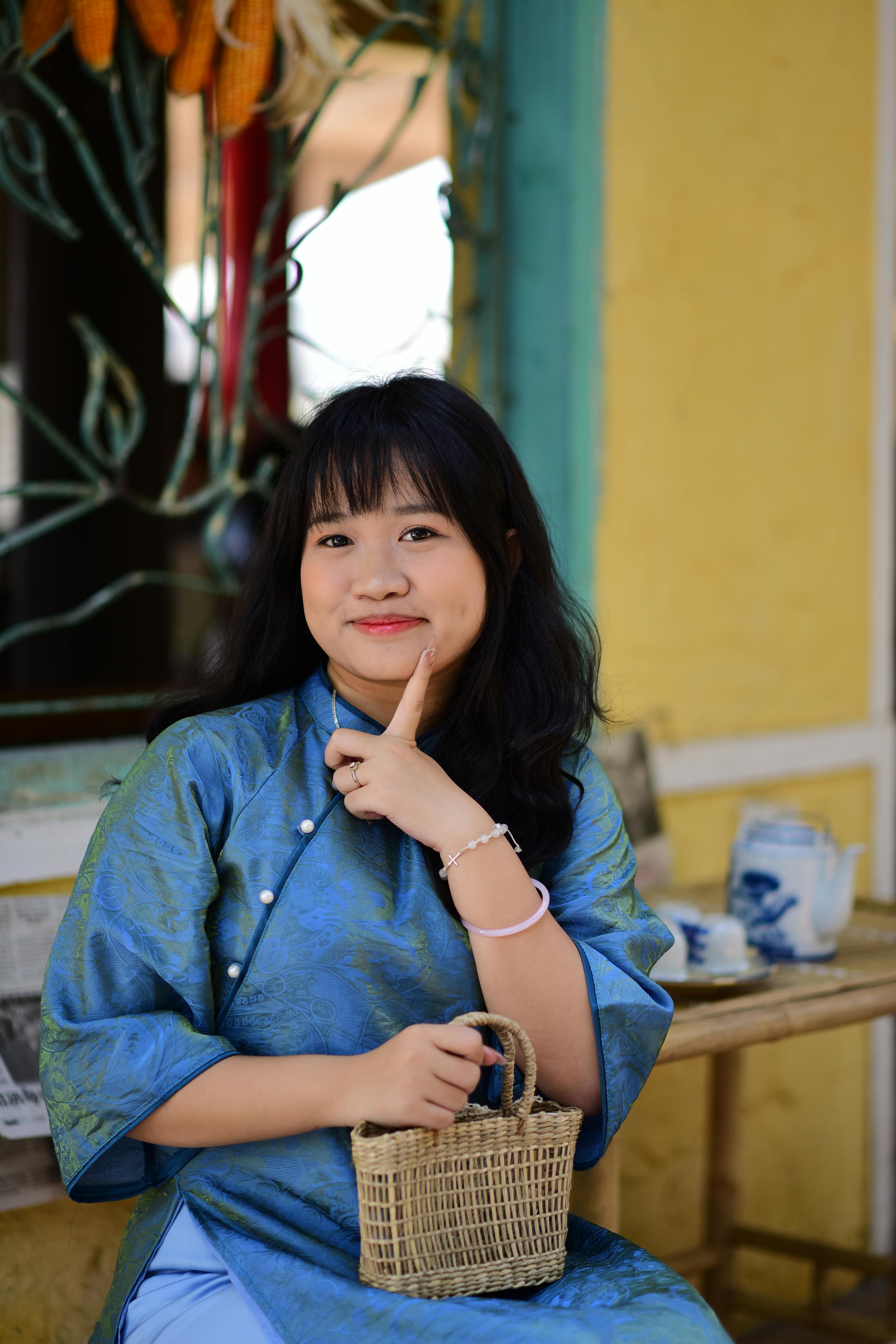 ฟรี หญิงสาวชาวเอเชียสวมชุดพื้นเมืองสีน้ำเงินนั่งอยู่กลางแจ้งพร้อมตะกร้าใบหนึ่งและกำลังยิ้ม คลังภาพถ่าย