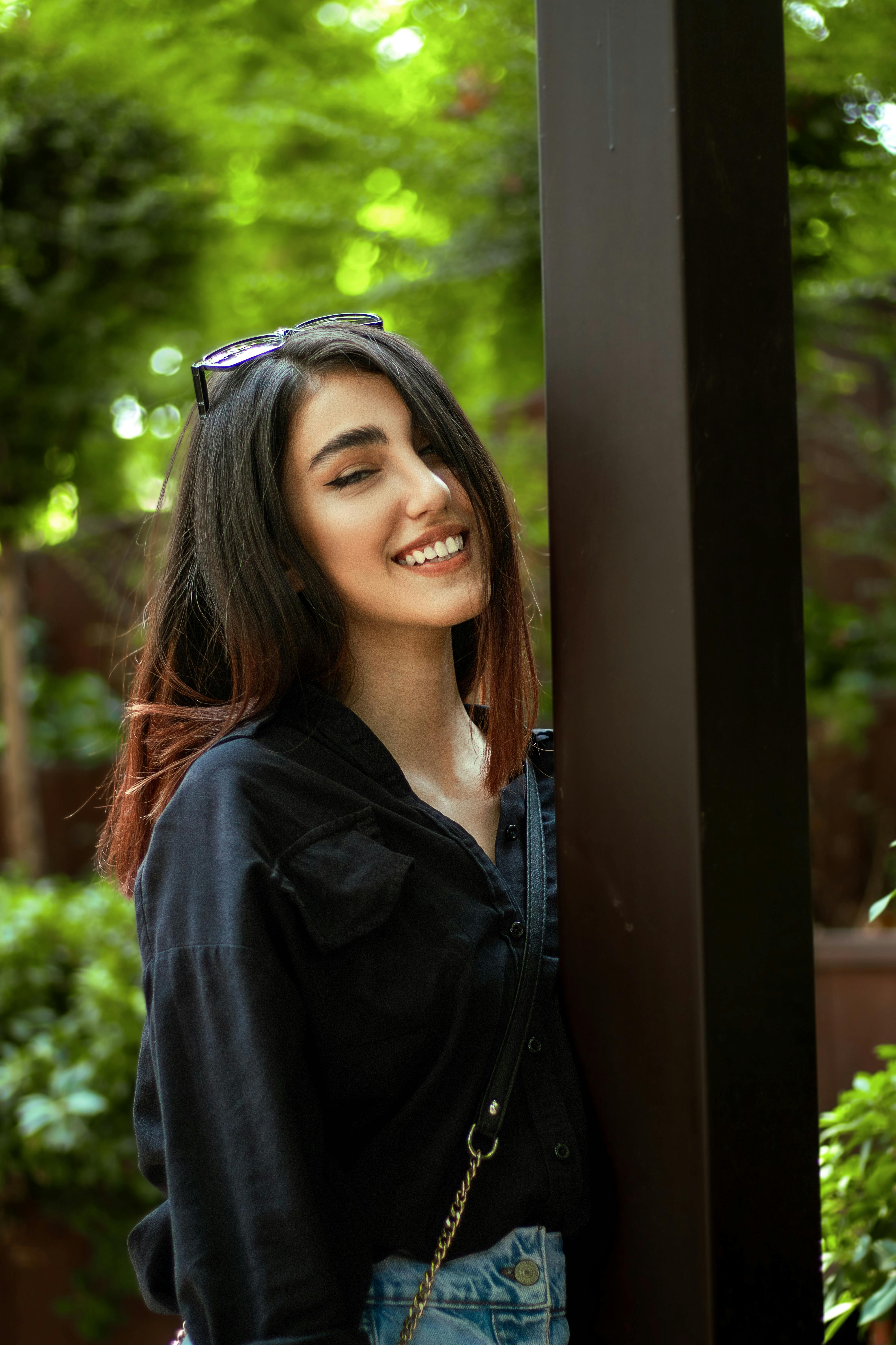 ฟรี หญิงสาวคนหนึ่งยิ้มอย่างอบอุ่นขณะพิงเสาอยู่ด้านนอก คลังภาพถ่าย