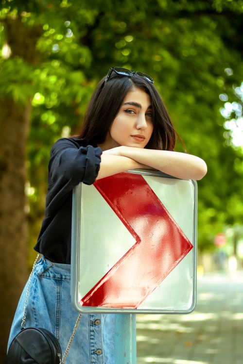 Free A young woman leans on a reflective road sign surrounded by lush greenery on a sunny day. Stock Photo