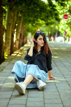 Stylish young woman in casual attire sitting on a tree-lined sidewalk during the day.