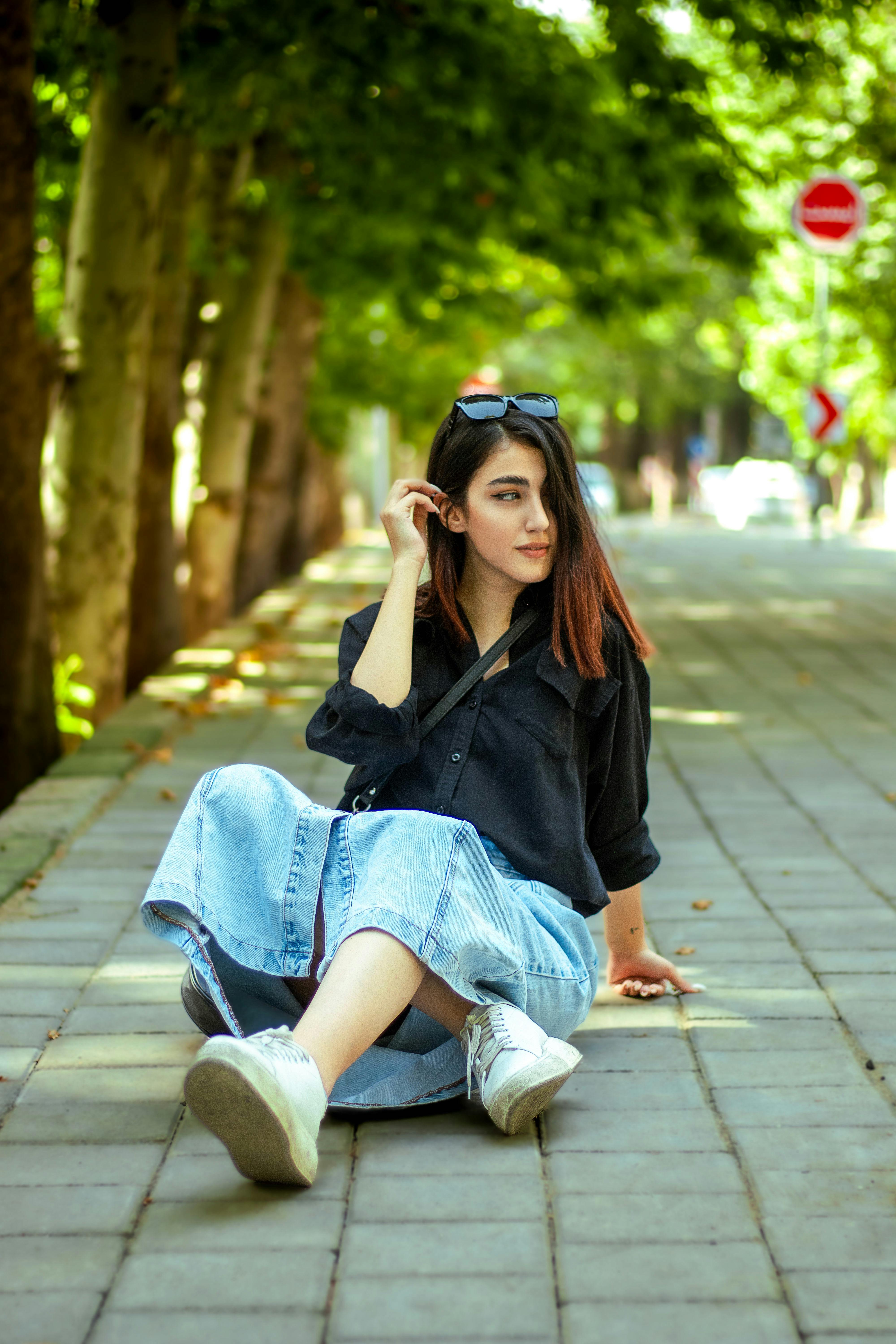 Free Stylish young woman in casual attire sitting on a tree-lined sidewalk during the day. Stock Photo
