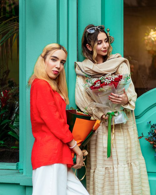 Free Two fashionable women holding flower bouquets pose by a teal doorway, showcasing modern style. Stock Photo