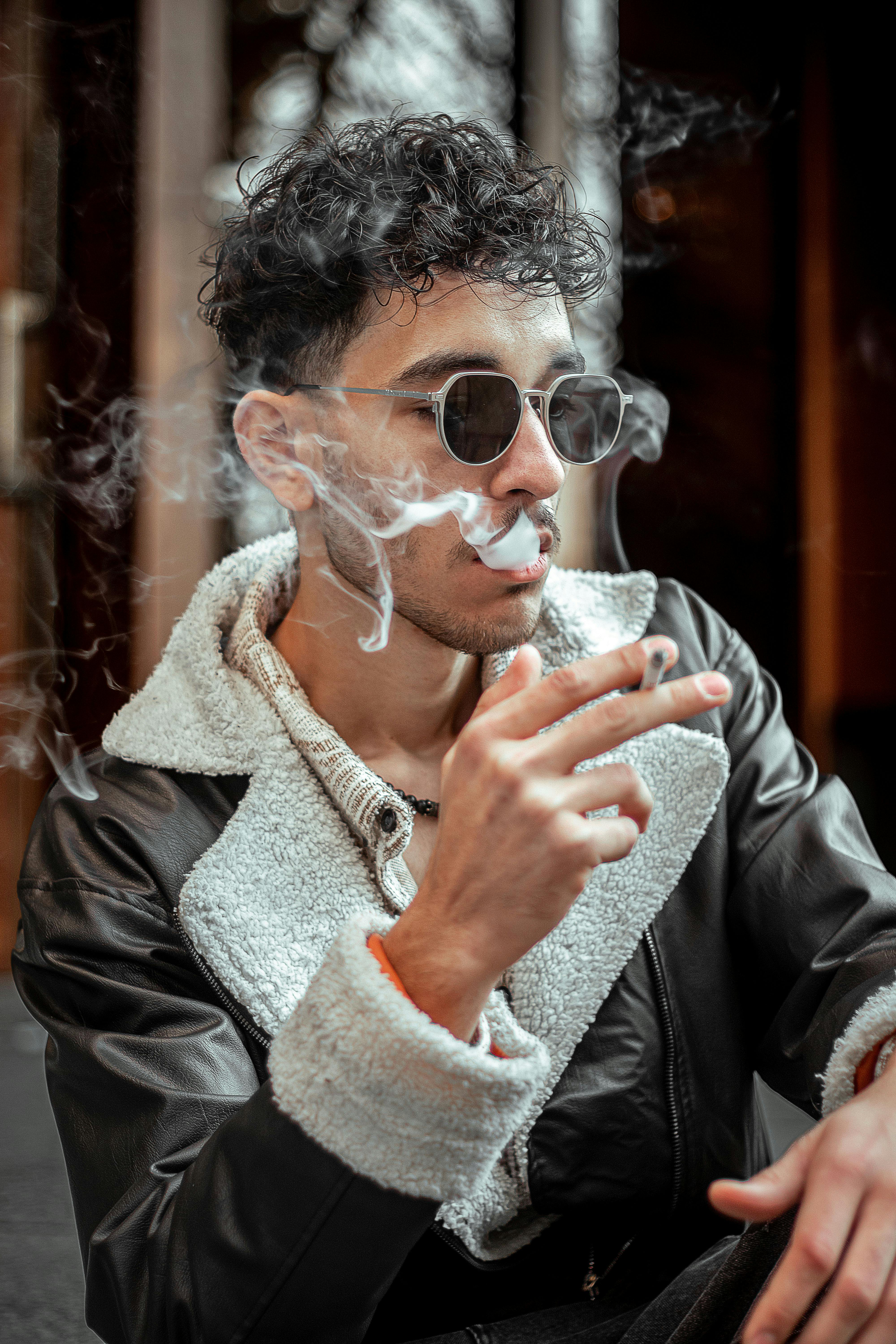 gratis Een jonge man met zonnebril en jas aan, rokend, met een stedelijke achtergrond. Stockfoto