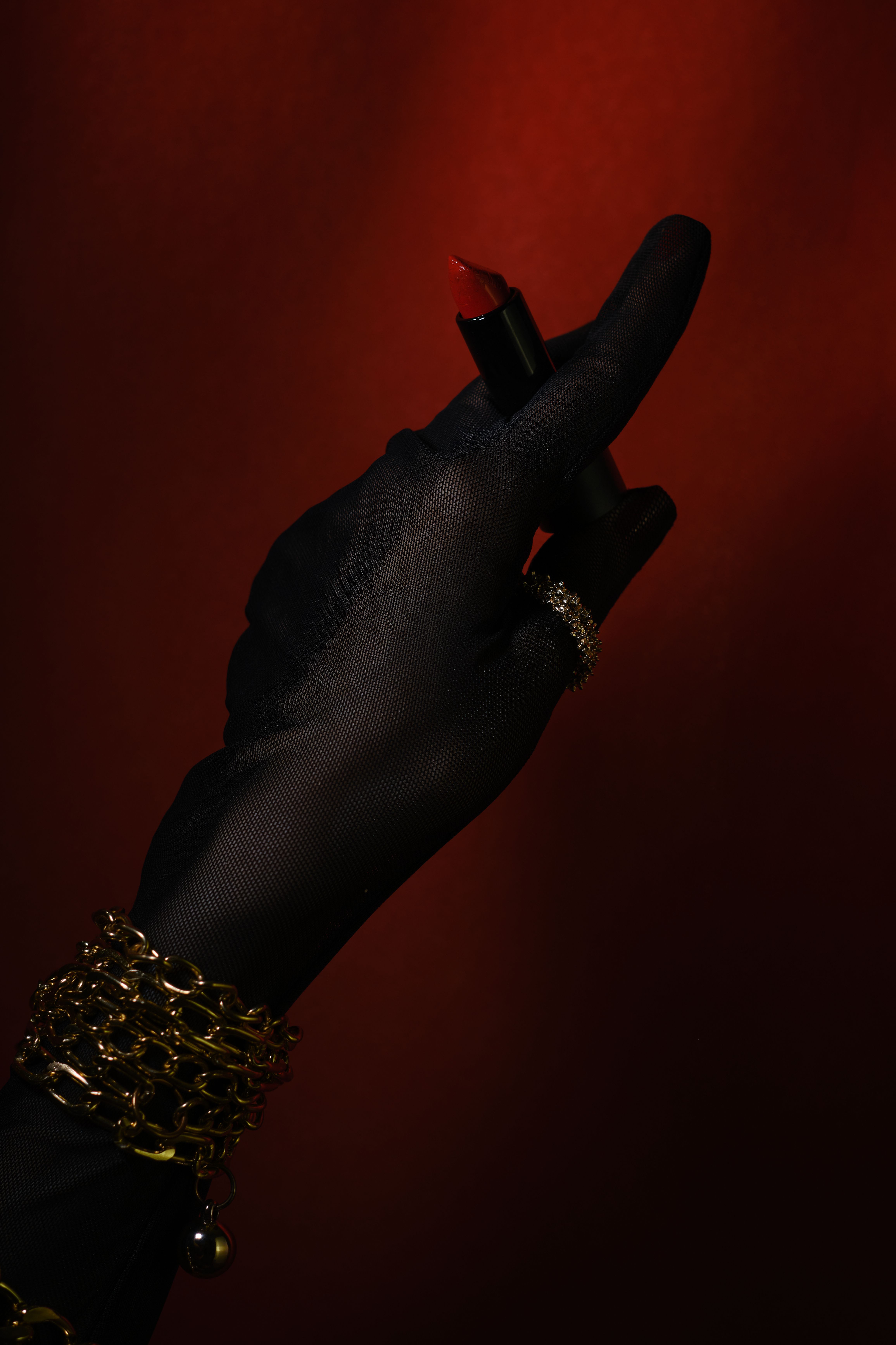 A stylish, black gloved hand displaying a luxurious red lipstick against a dark red background.
