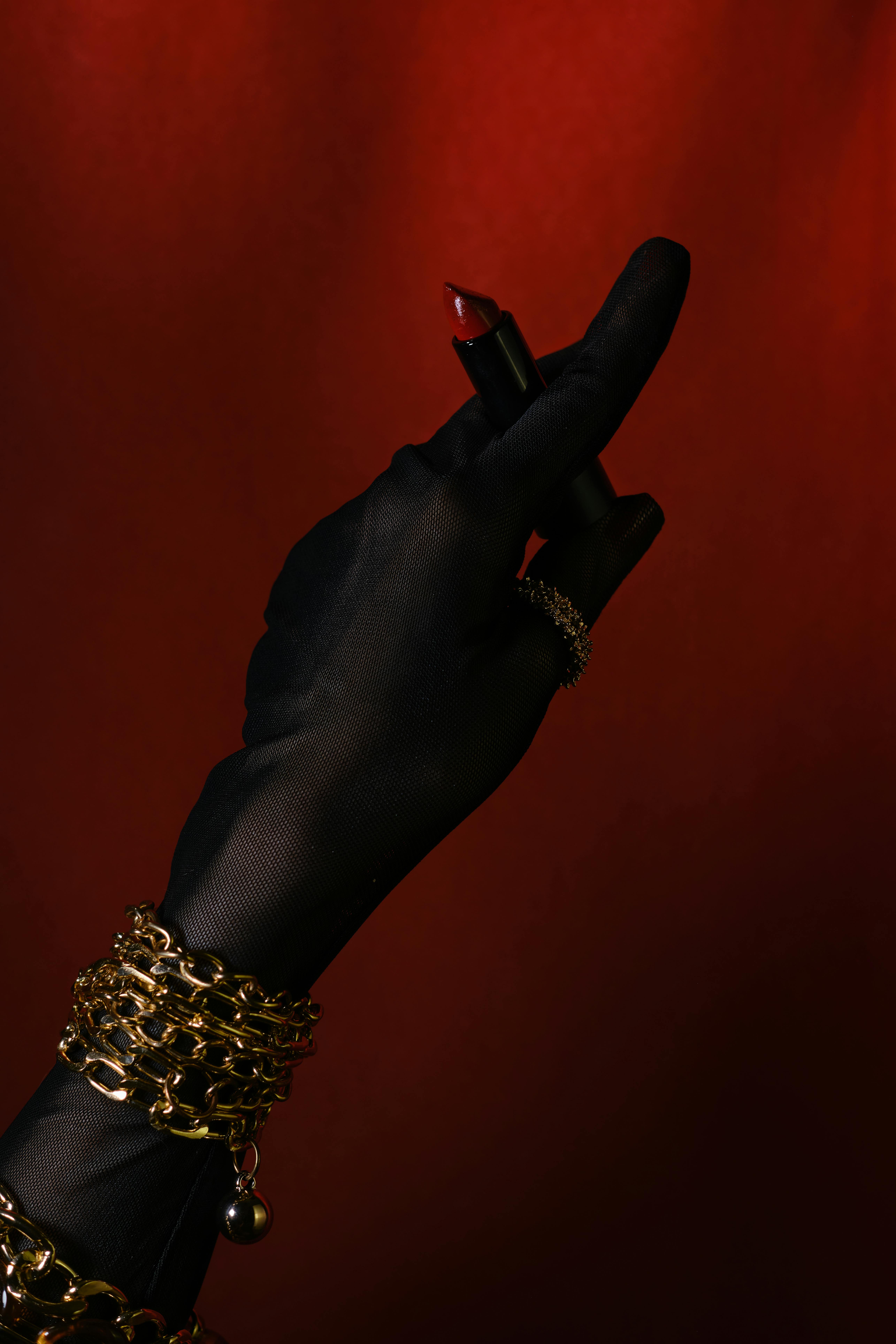 Gratuit Mână înmănușată neagră care ține ruj roșu pe un fundal roșu intens, accesorizată cu bijuterii de aur. Fotografie de stoc