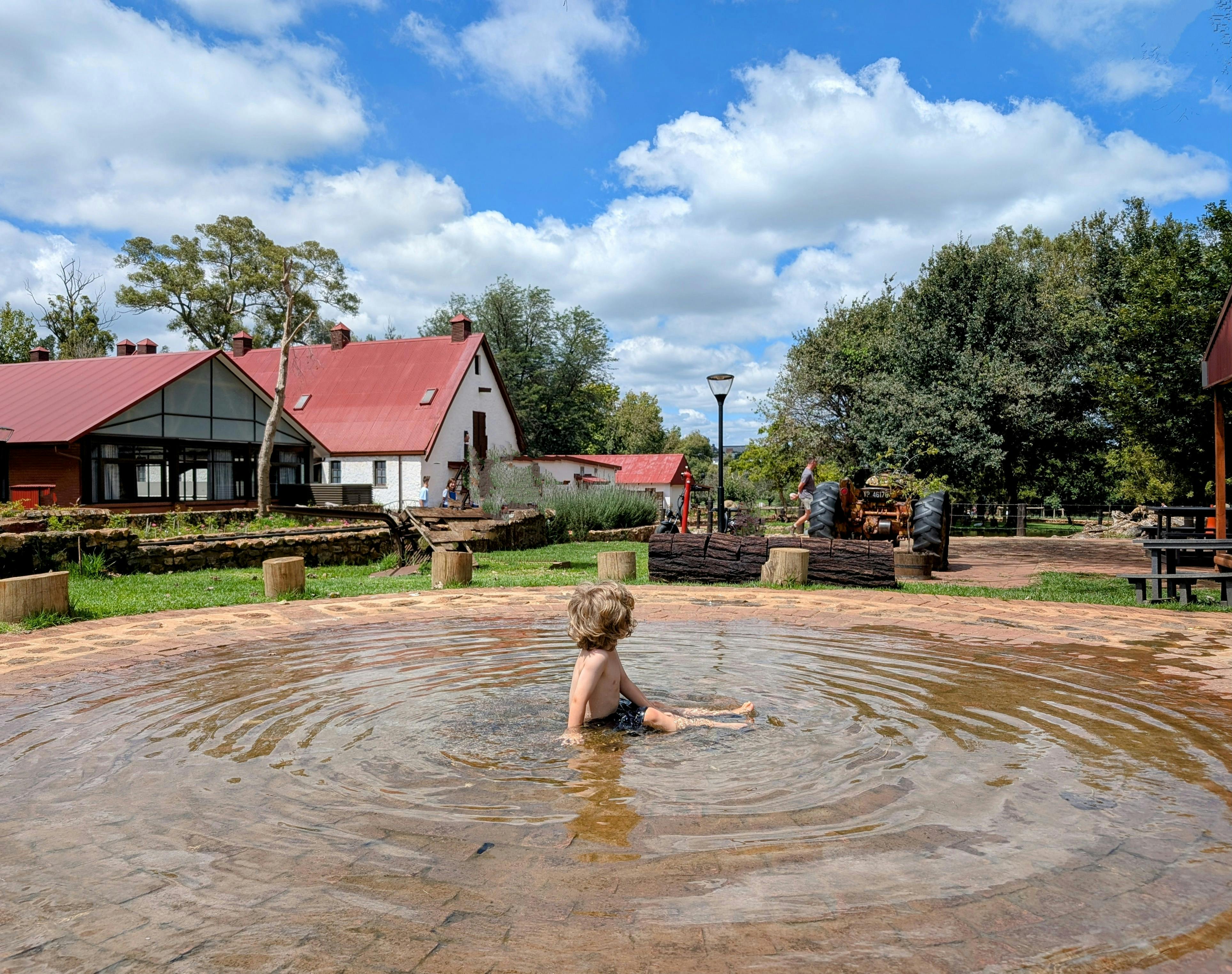 Miễn phí Một em bé đang thích thú nghịch nước trong bể bơi ngoài trời gần những ngôi nhà mộc mạc. Ảnh lưu trữ