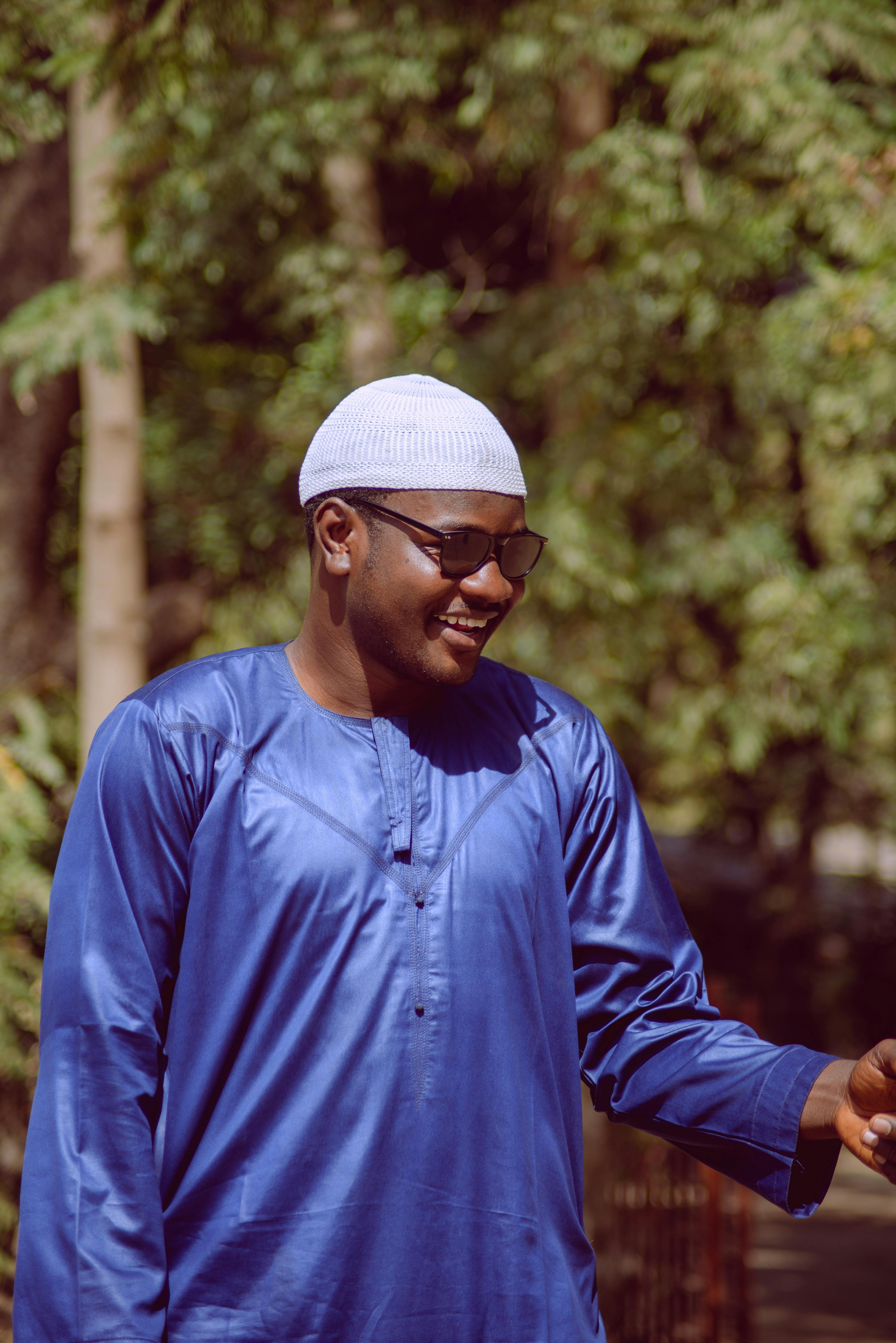 A happy man in blue traditional attire and a white cap outdoors in Zaria, Nigeria.