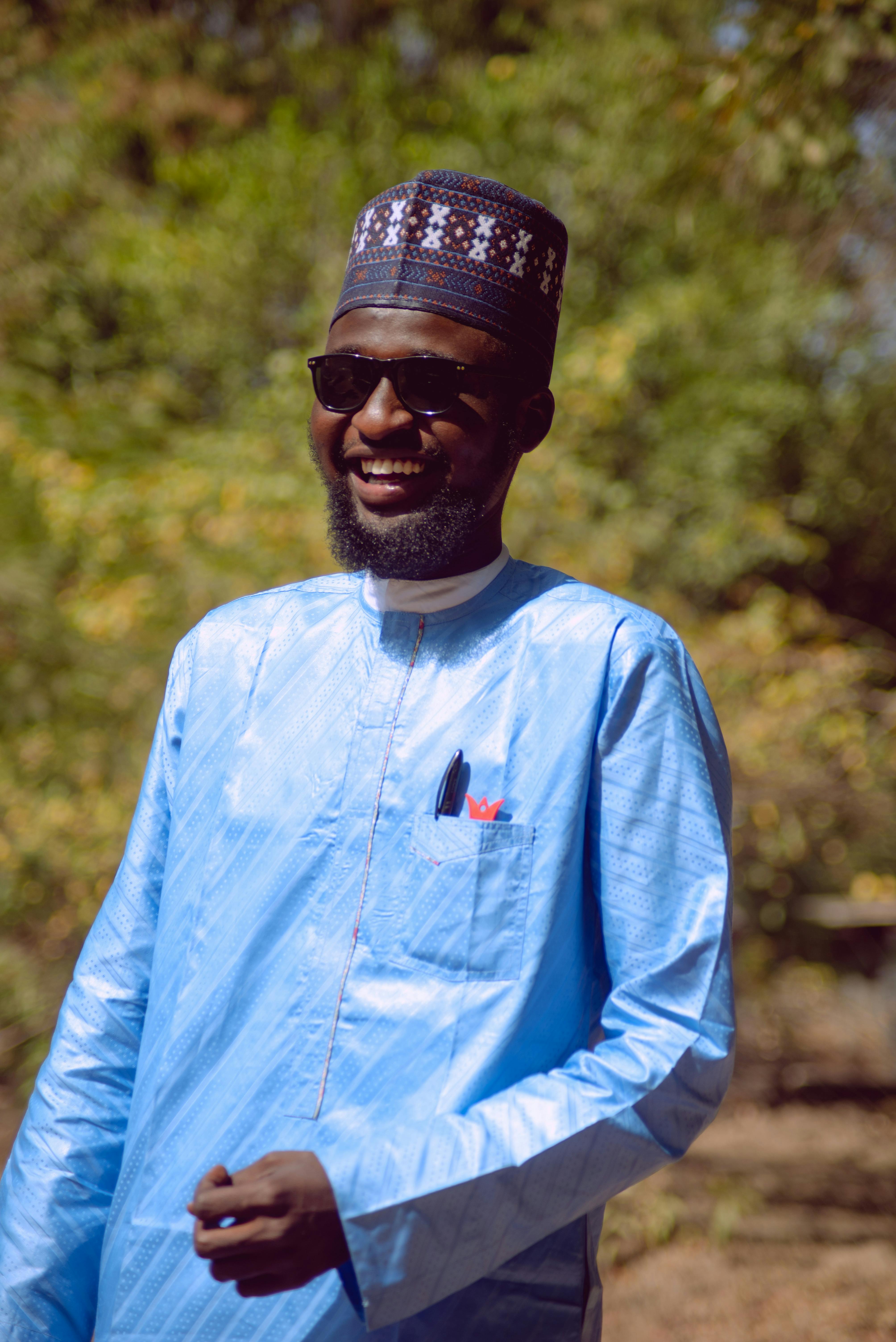 A man in traditional Nigerian attire smiles outdoors in Zaria, Nigeria, under bright sunlight.