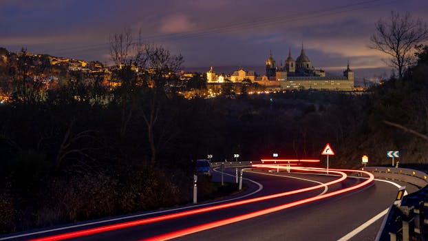 Night view of El Escorial with light trails on winding road, capturing a dramatic cityscape scene.