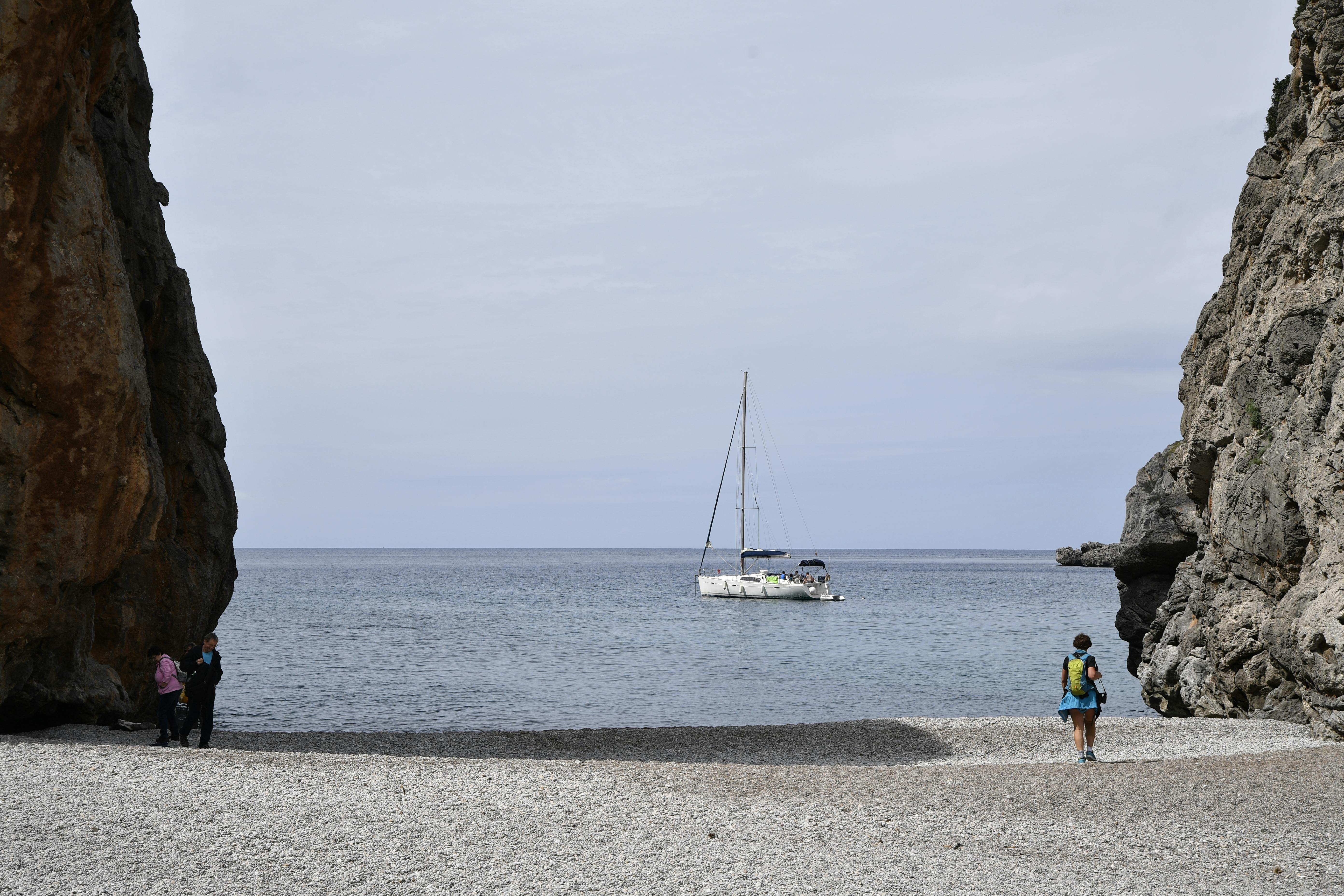 Δωρεάν Γαλήνια παραλία με βότσαλα με γκρεμούς και ιστιοπλοϊκό στις Βαλεαρίδες Νήσους, Ισπανία. Φωτογραφία από στοκ φωτογραφιών