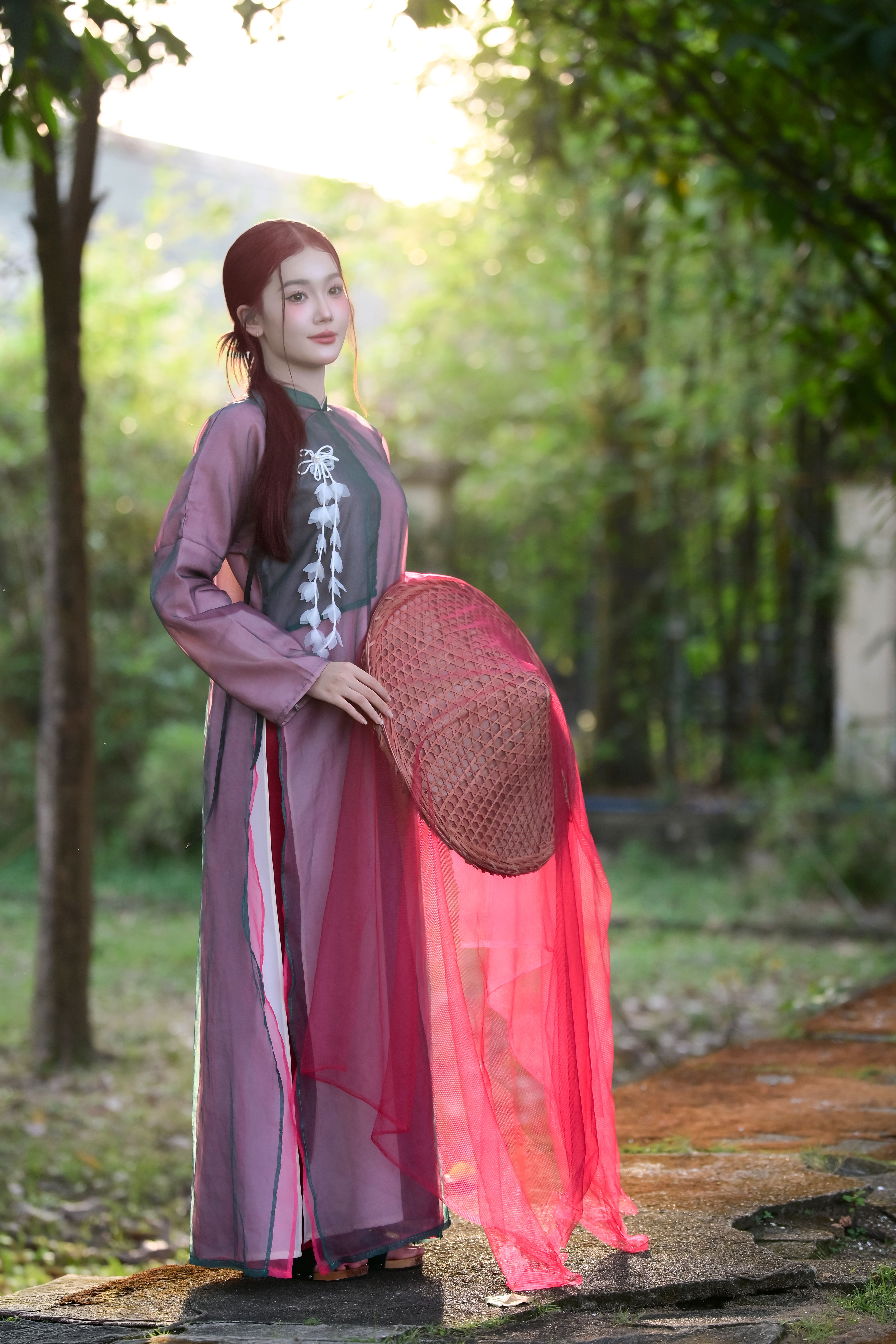 A woman in traditional Vietnamese attire holding a hat, set against a serene outdoor backdrop with warm lighting.