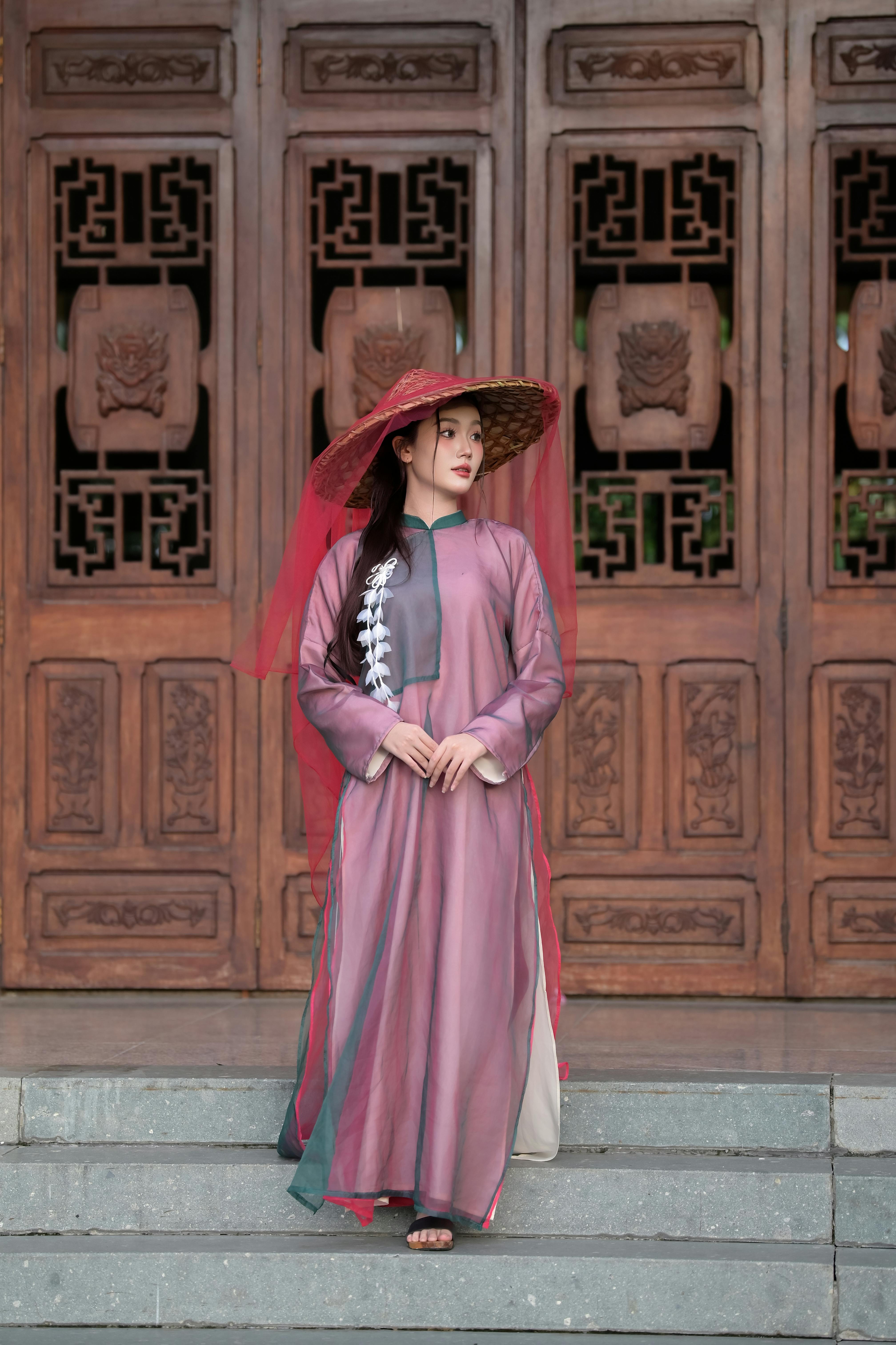 Woman in traditional Asian dress and hat standing by ornate wooden doors.