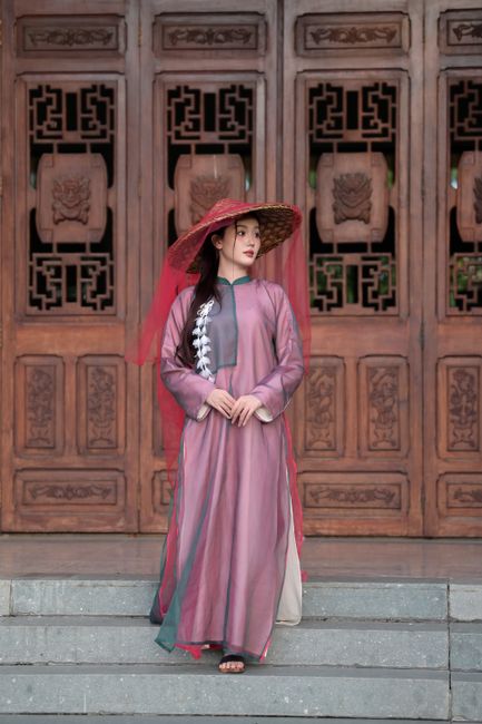 Woman in traditional Asian dress and hat standing by ornate wooden doors.