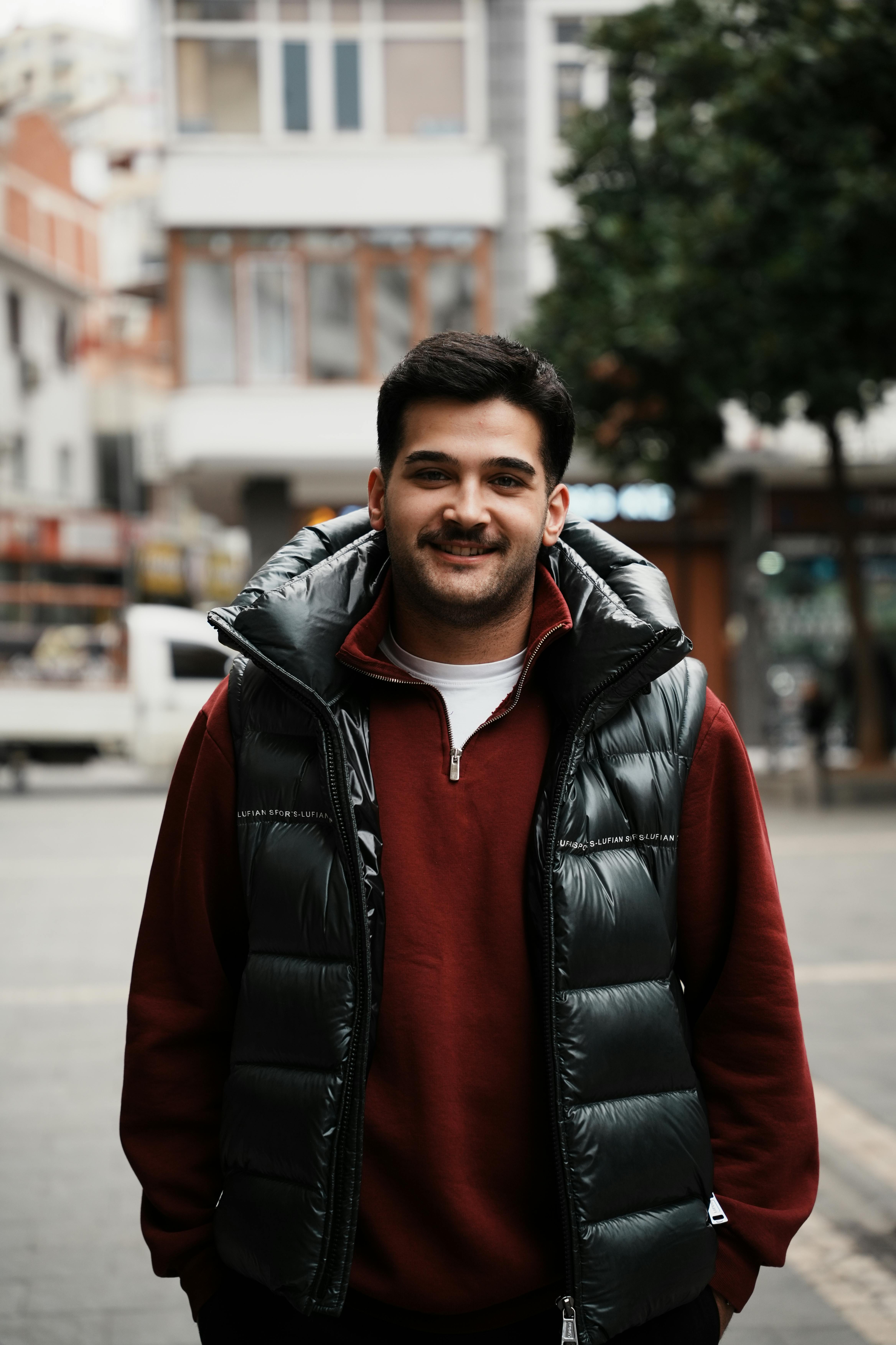 Gratis Un uomo sorridente con un maglione rosso e un piumino è in piedi all'aperto in una strada cittadina. Foto a disposizione