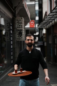 Bearded man in a black sweater serves traditional Turkish tea on a city street.