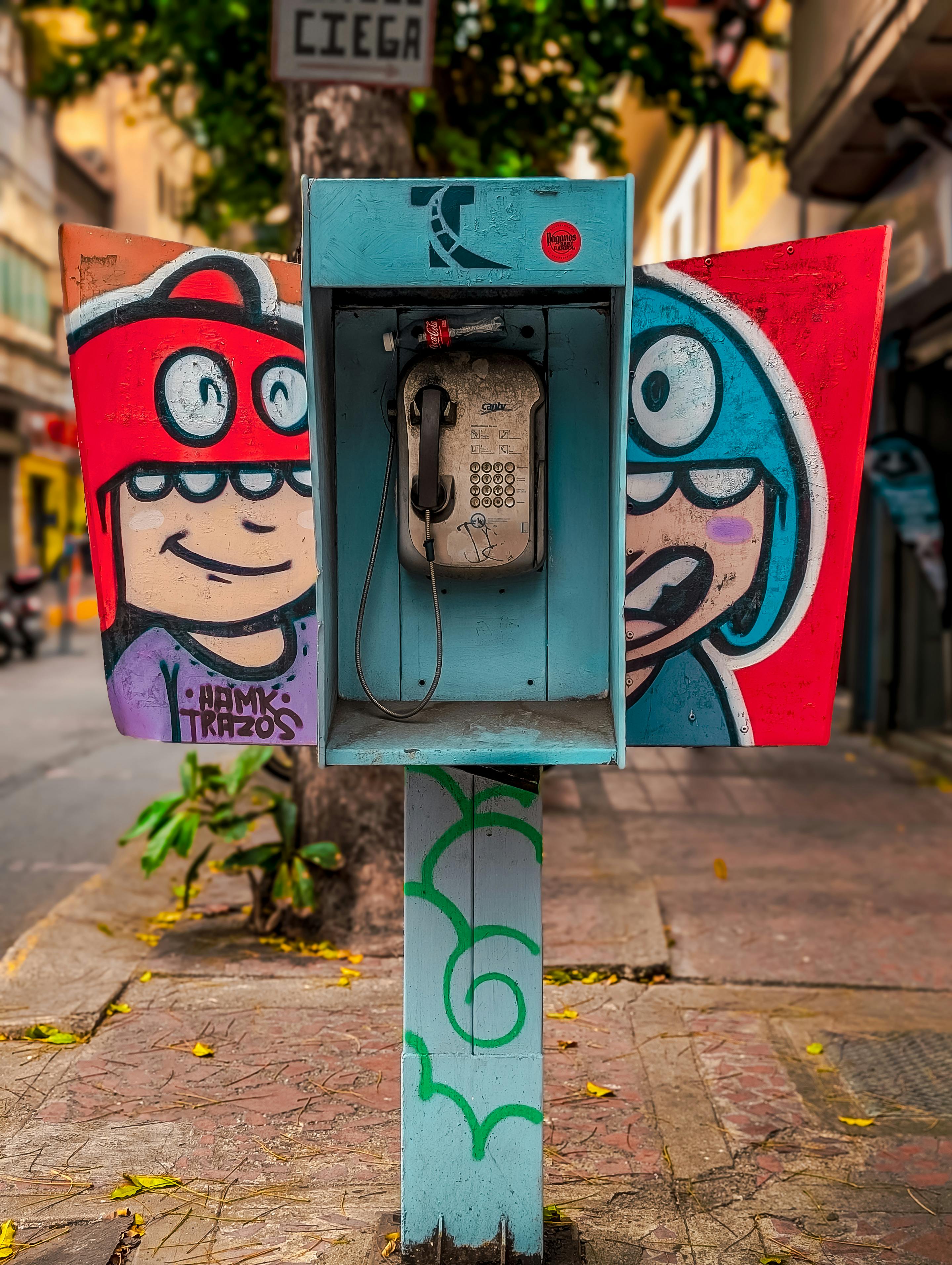Bezpłatne Żywa sztuka uliczna na zabytkowej budce telefonicznej przedstawiająca rysunkowe twarze na tle miejskiego otoczenia. Zdjęcie z galerii