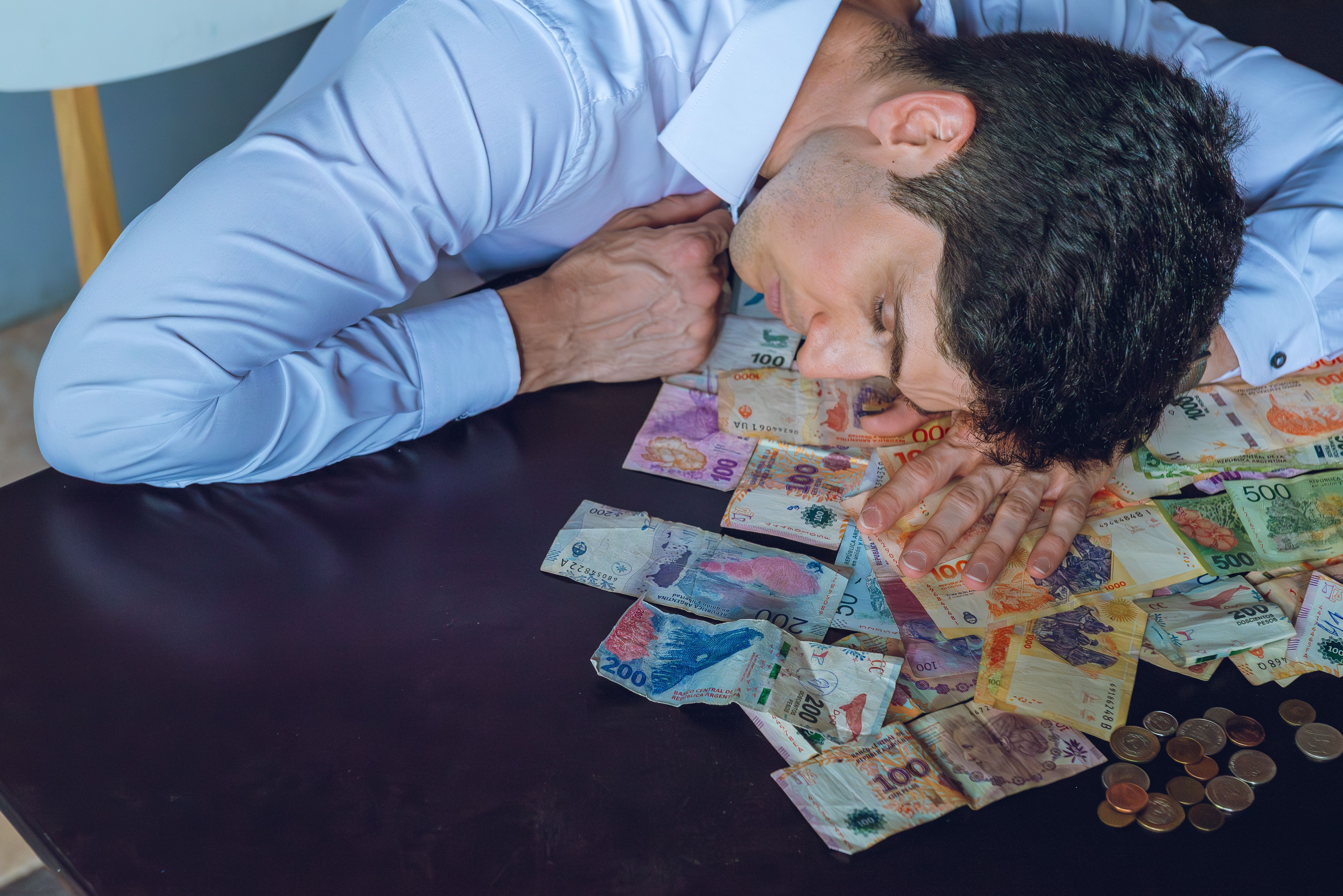 A tired man rests on a table covered with Argentine pesos, symbolizing financial stress.