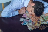 A tired man rests on a table covered with Argentine pesos, symbolizing financial stress.